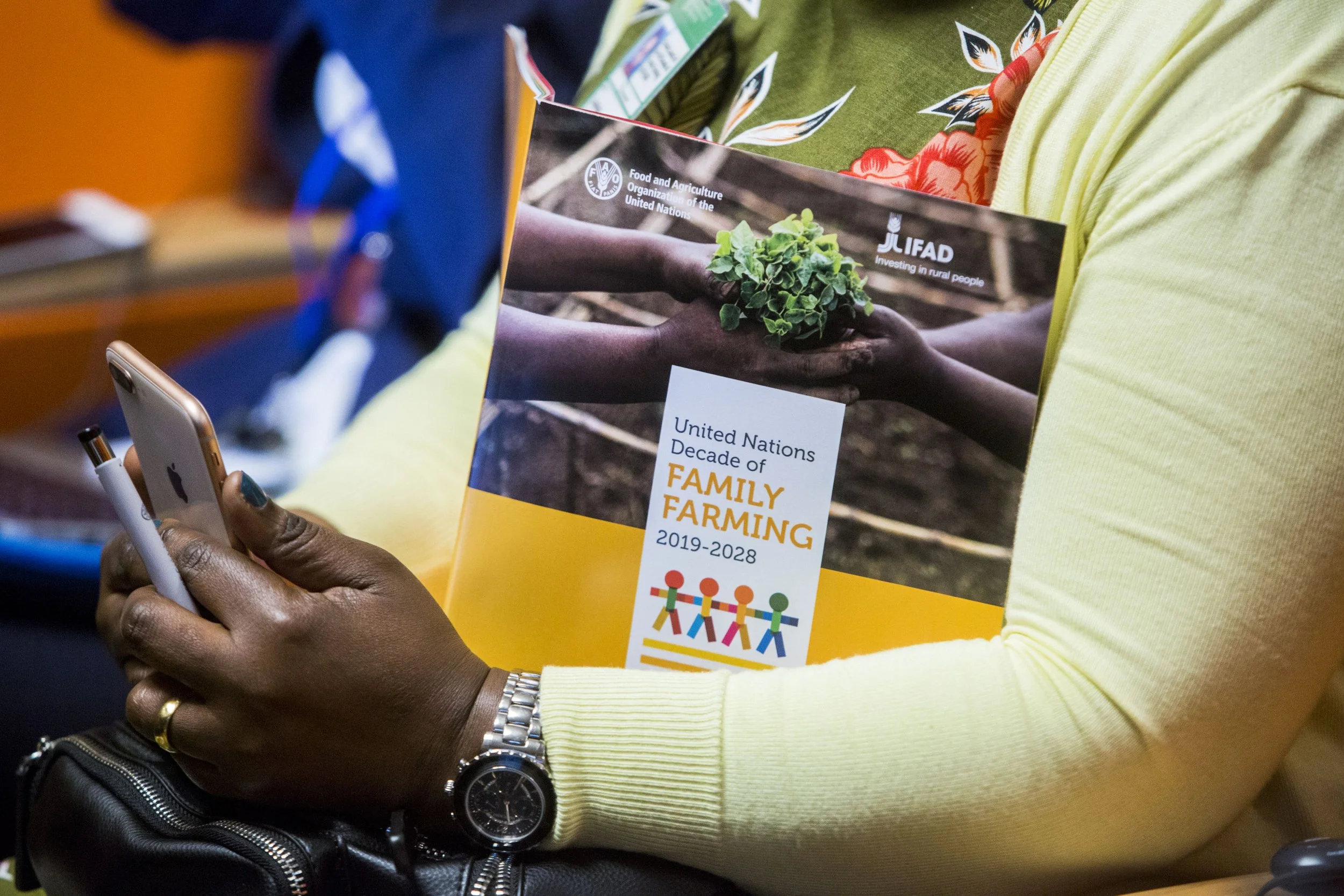 A person holding a pamphlet about family farming, with the United Nations logo and text, while using a smartphone. The person is wearing a yellow jacket, a silver watch, and a ring, with a black purse nearby.