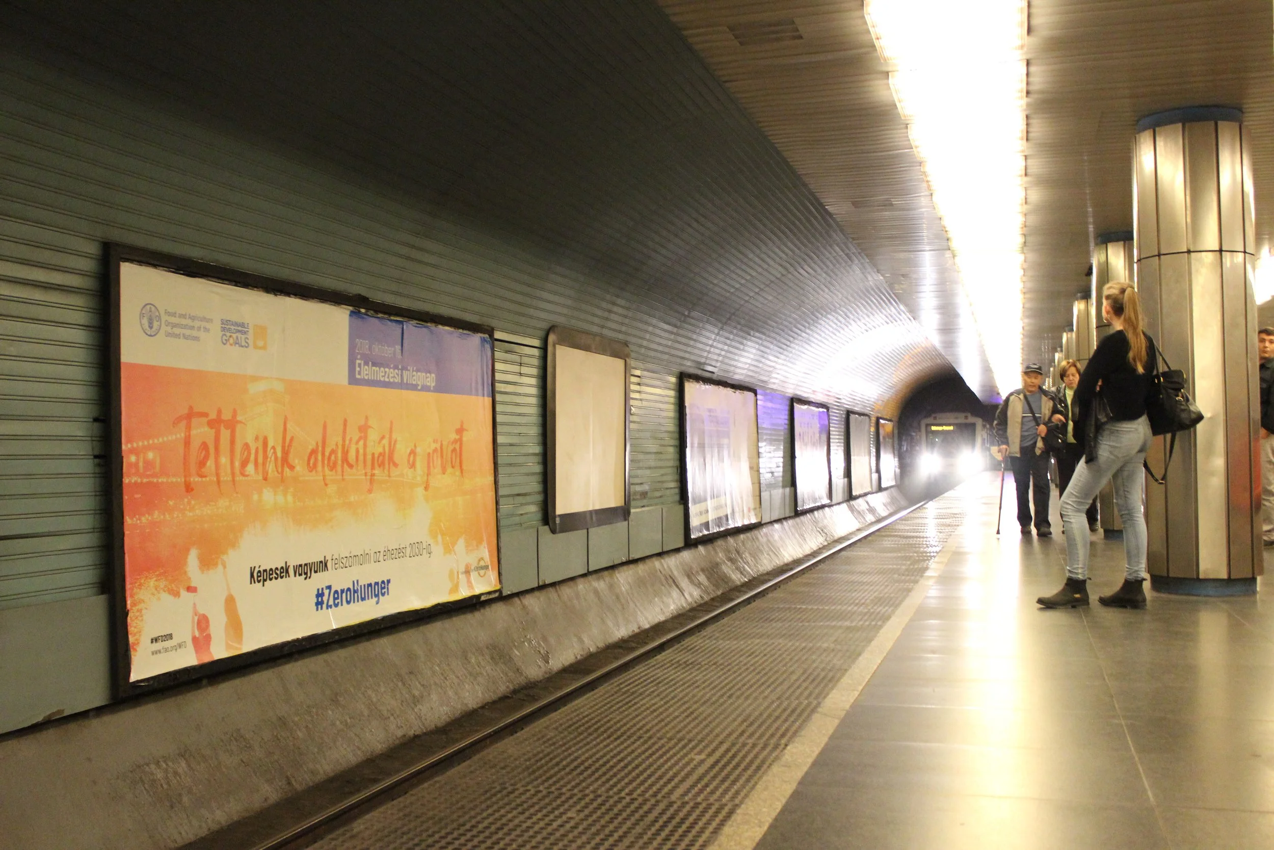 People waiting at an underground metro station, with advertisements on the wall and a train approaching in the distance.