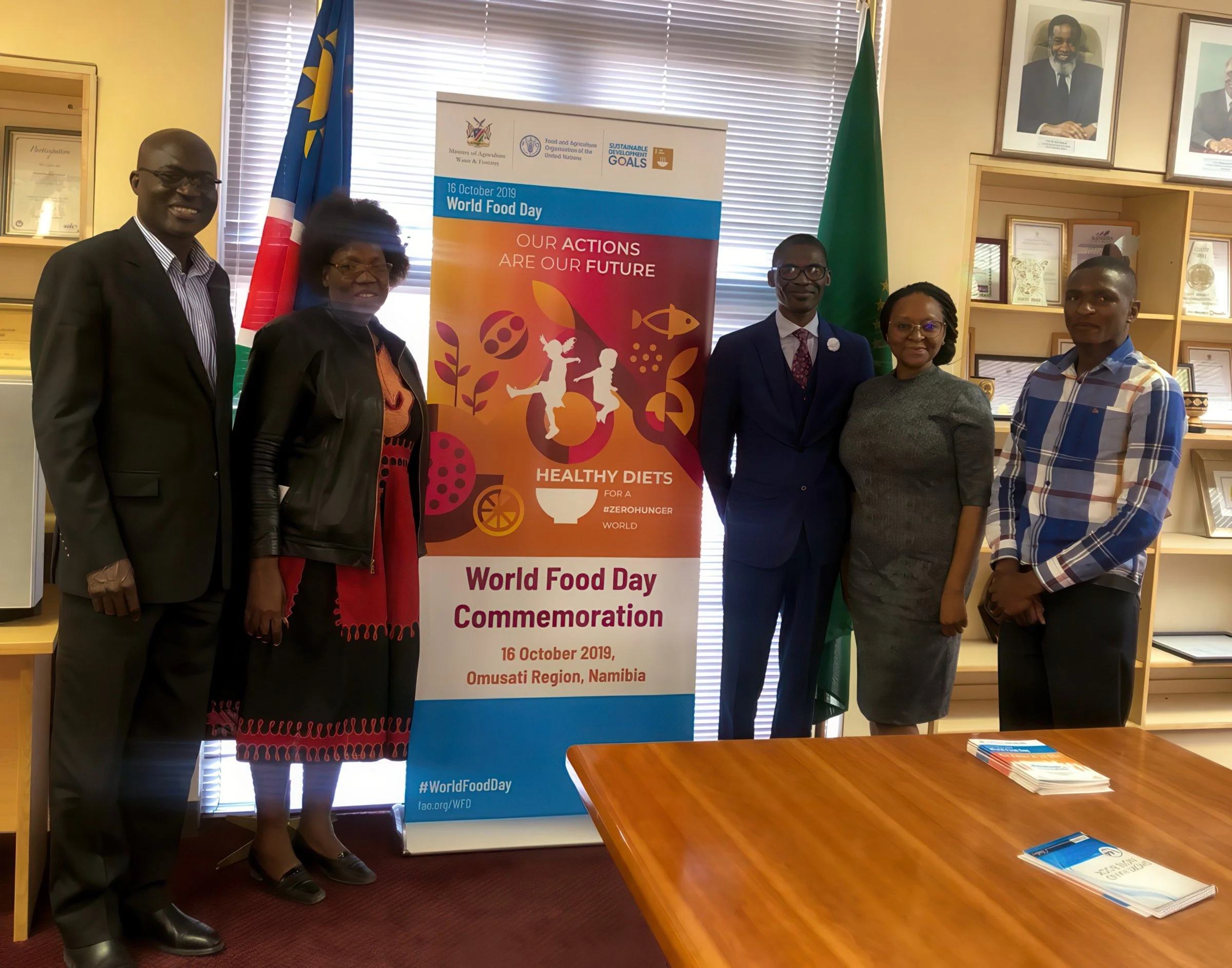 Group of six people standing in front of a World Food Day poster in an office. The poster mentions the date October 16, 2019, and the location Namibia. There are flags and framed pictures on the walls.