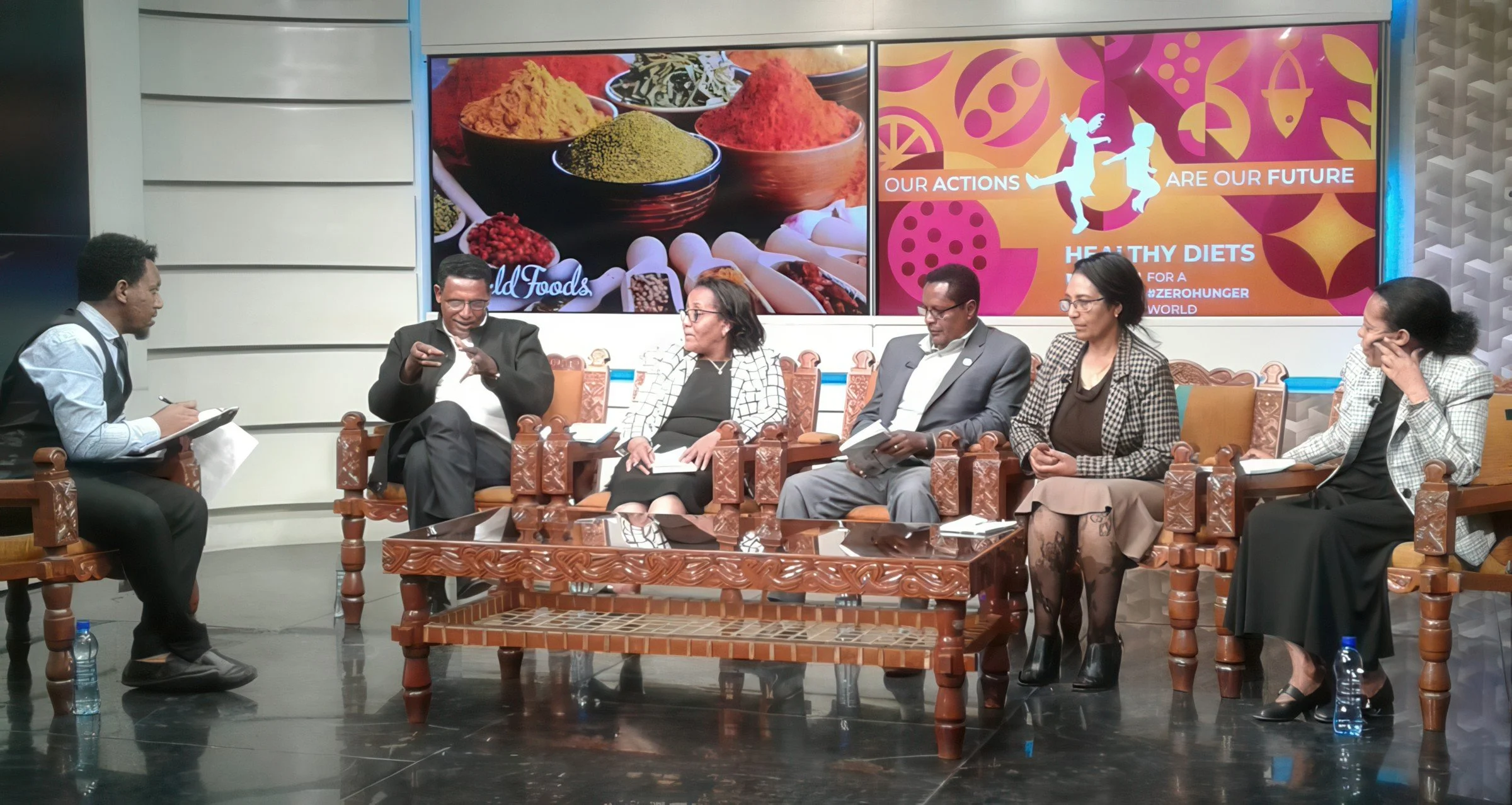 A panel of six people seated in front of a large screen displaying images of spices and food, during a discussion or presentation, with bottles of water on the floor nearby.