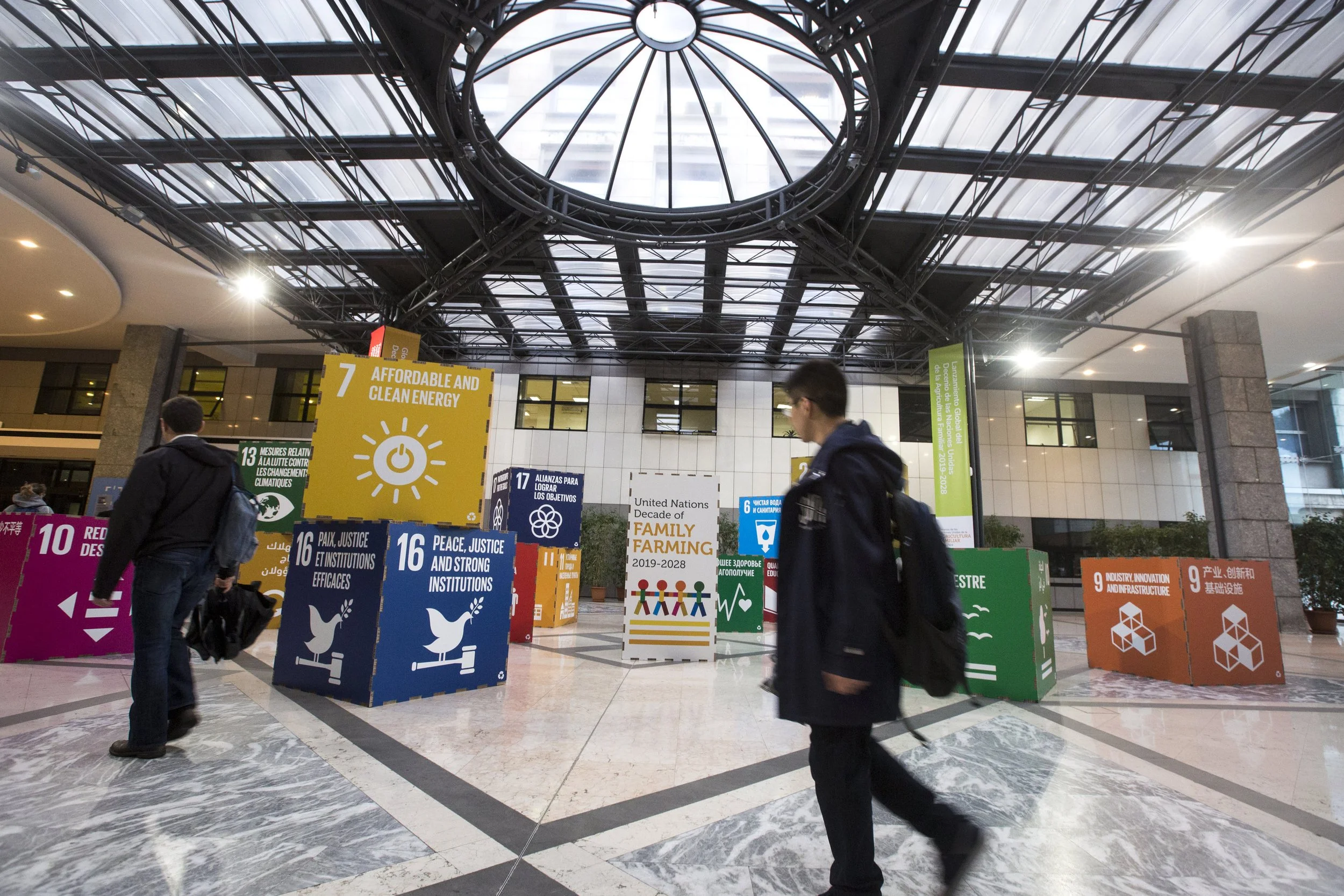 Interior view of a conference or exhibition hall with people walking past colorful banners promoting United Nations Sustainable Development Goals, including clean energy, climate measures, peace and justice, family farming, and infrastructure, under 