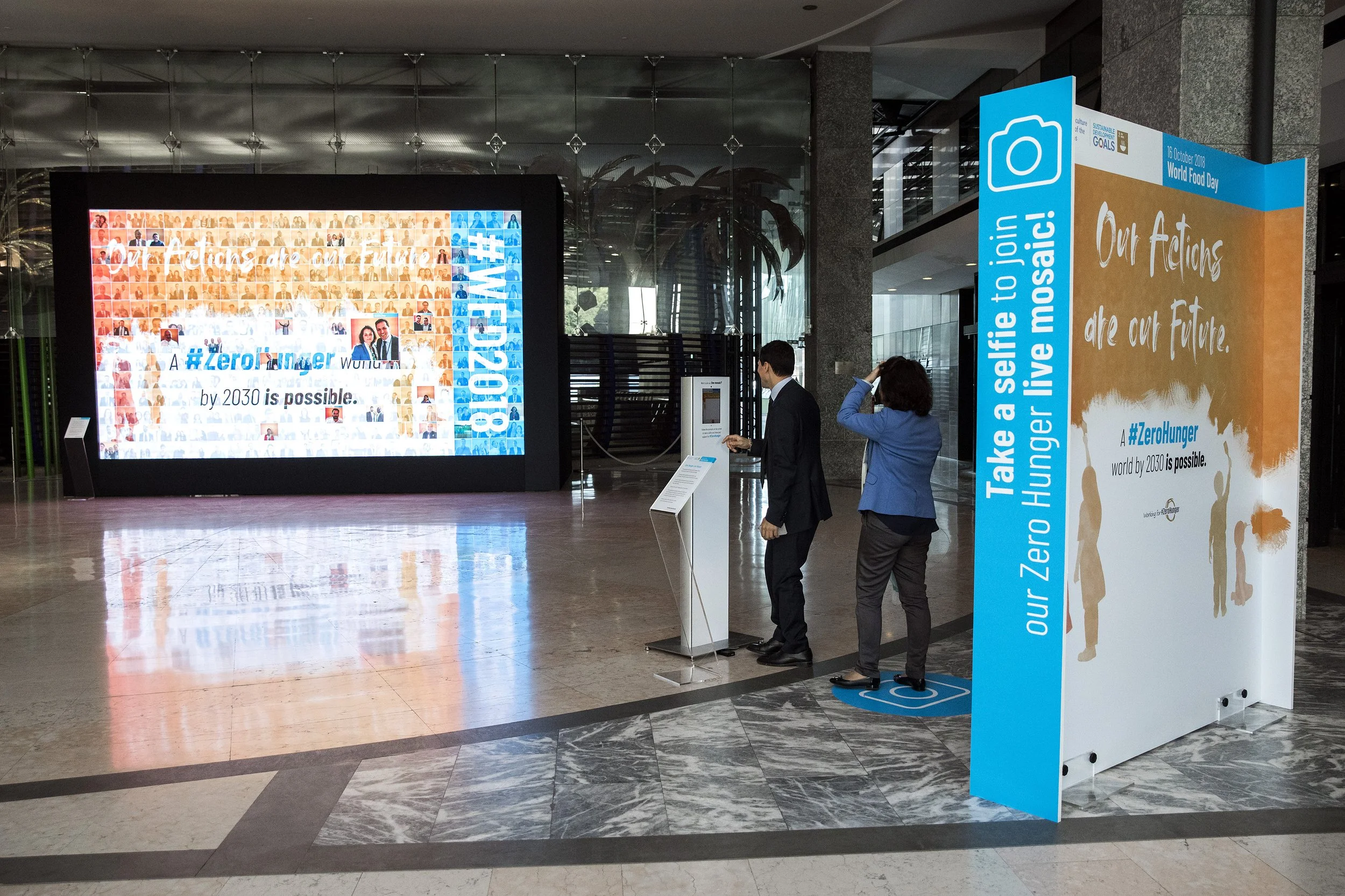 Indoor lobby area with digital screen and photo booth for World Food Day. The screen displays a collage of photos and text reading 'Zero Hunger by 2030 is possible.' Two people are using the photo booth, which features graphics and text about ending 
