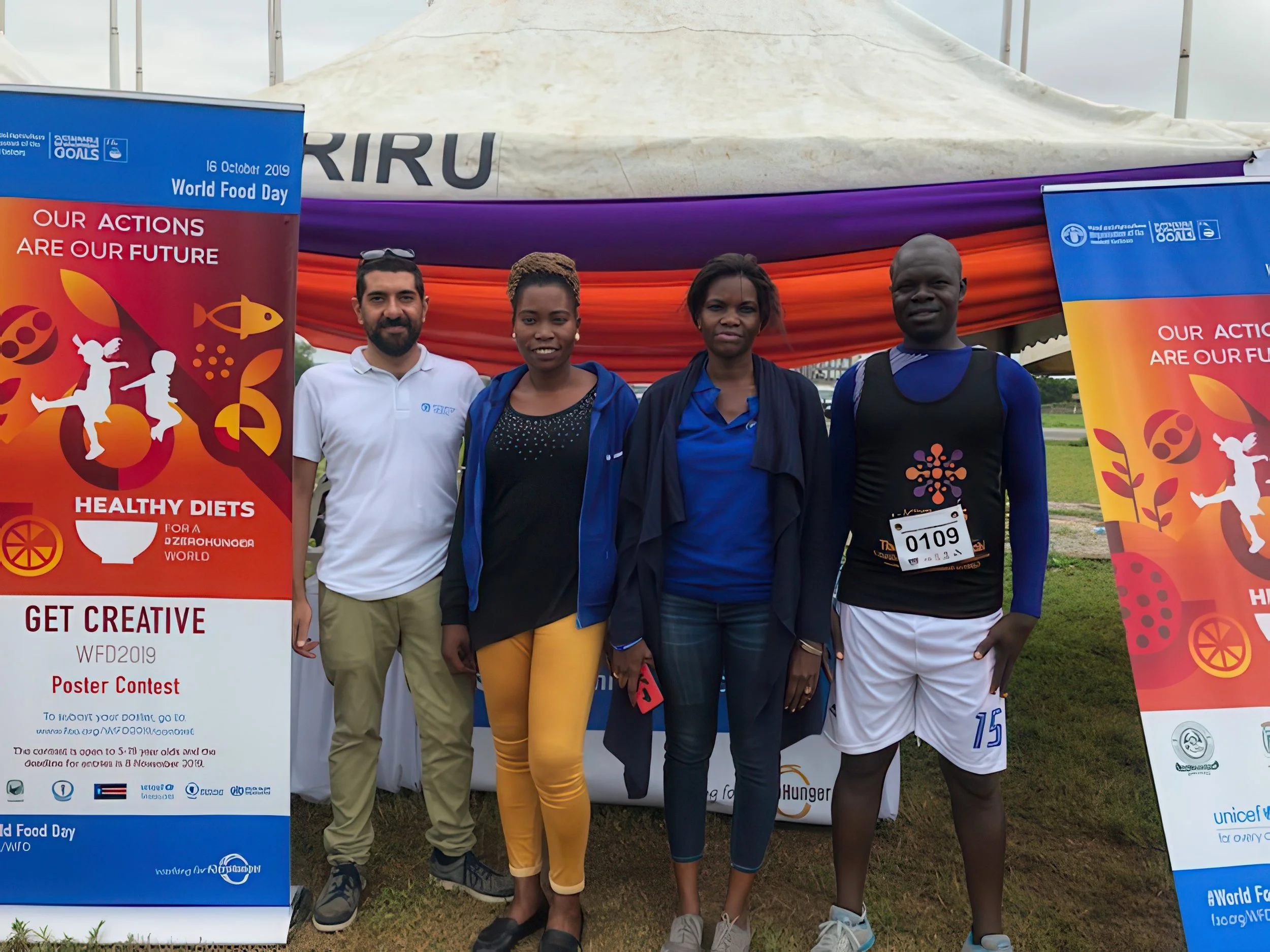 A group of four people standing outdoors in front of banners and a tent, participating in a World Food Day event, with one person wearing a race bib with the number 0109.