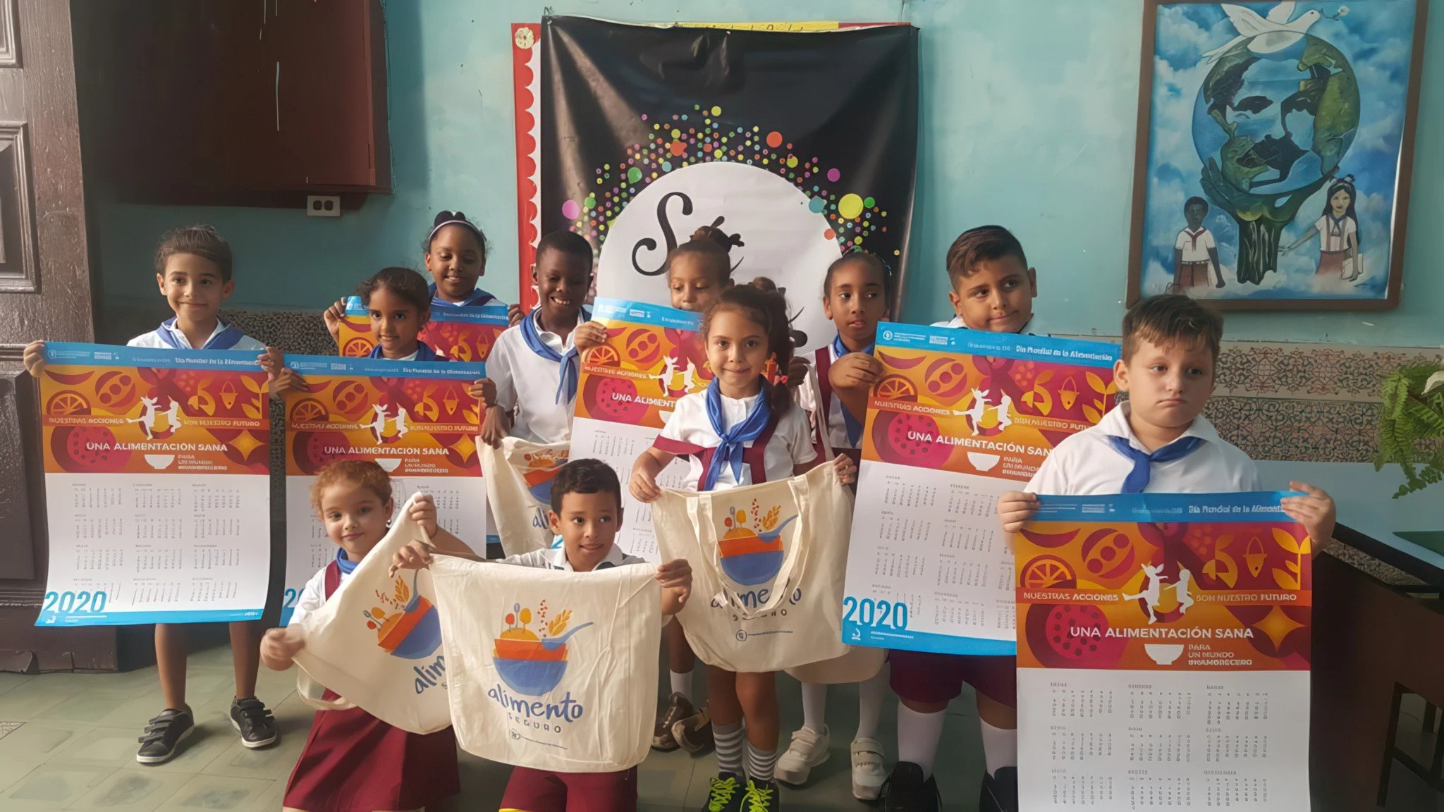Group of children in school uniforms holding posters and tote bags at an event promoting healthy eating in 2020, with classroom decorations and a large World Earth painting in the background.