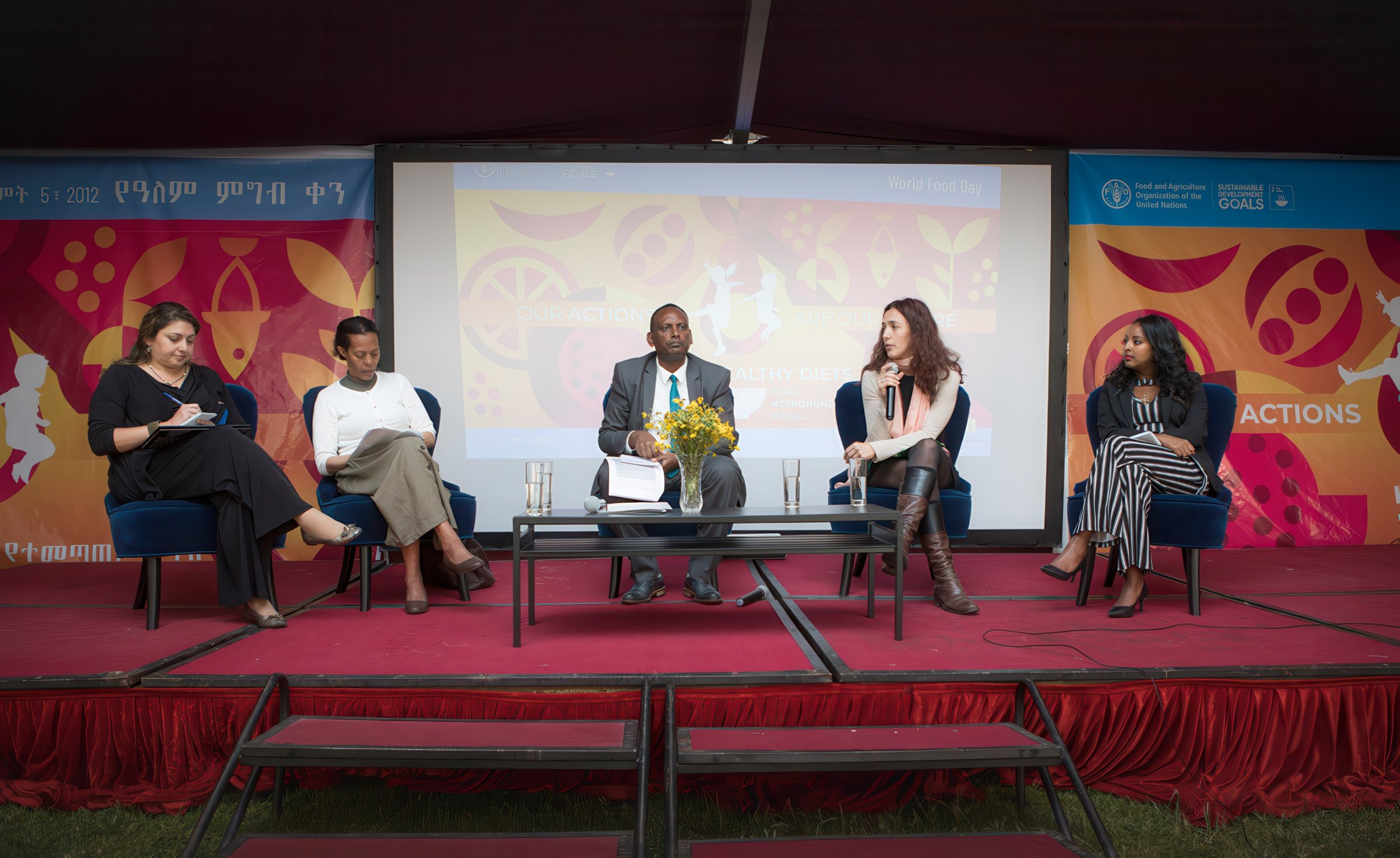 A panel discussion with five women and one man seated on stage, with a colorful backdrop and a large screen displaying 'World Food Day' and related graphics.
