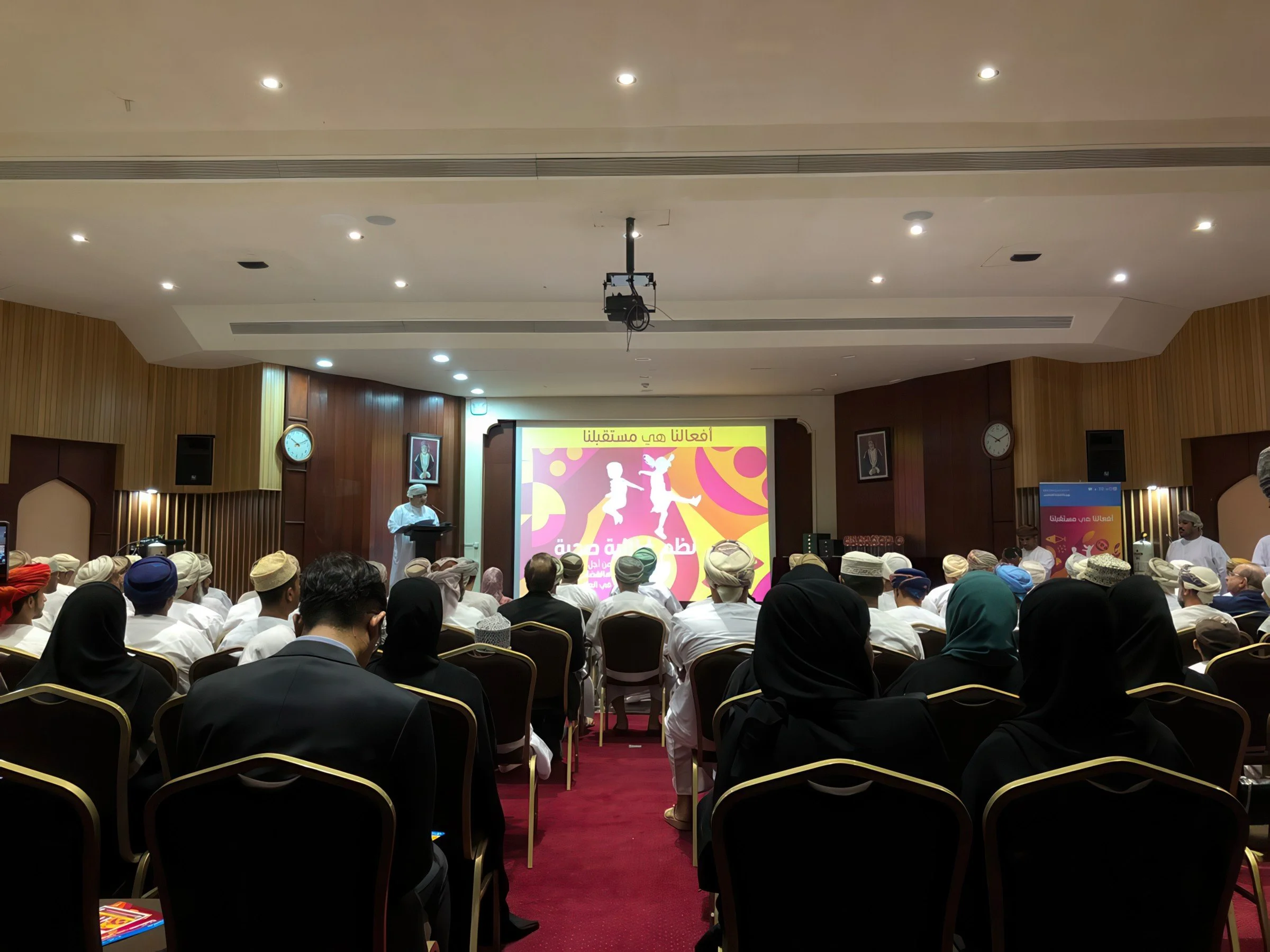 A large group of people seated in rows inside a conference room, facing a stage with a colorful presentation screen, with some individuals wearing traditional caps and headscarves, and a clock on the wall showing the time as approximately 8:15.