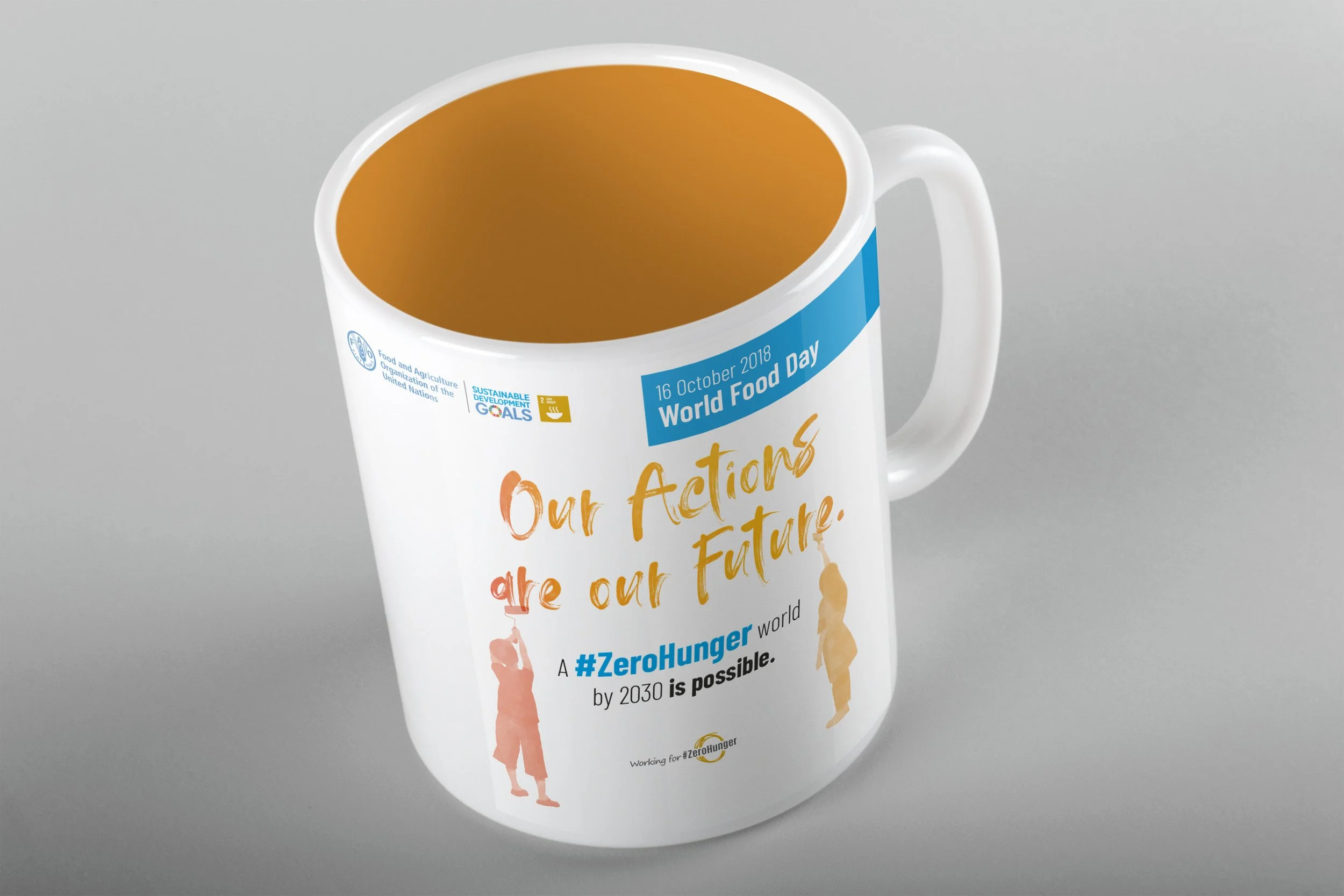 White mug with blue band around top featuring logos and text about World Food Day; orange liquid inside; part of the mug's message visible reading 'Our actions are our future' and promoting #ZeroHunger by 2030.