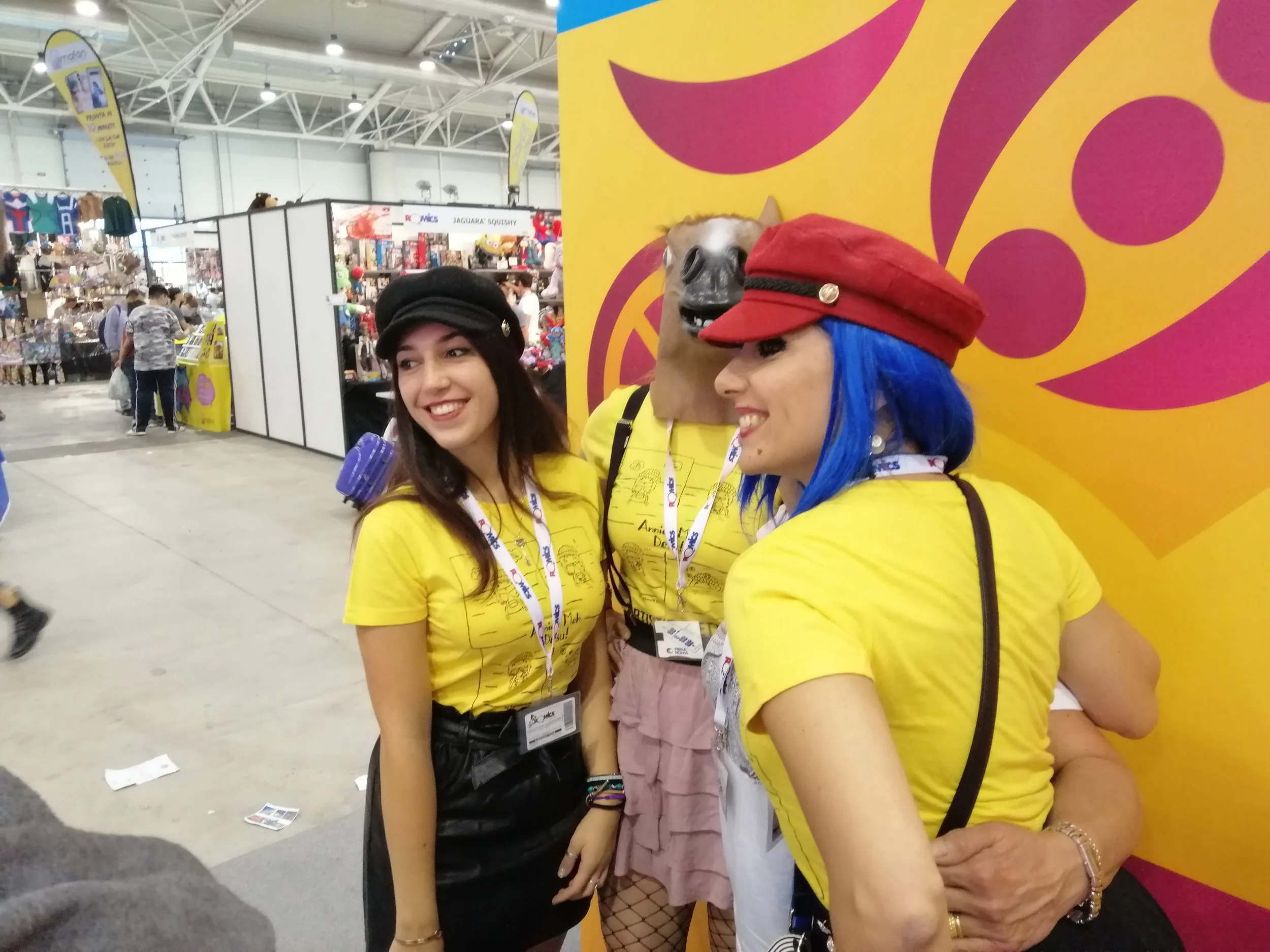 Three women wearing yellow shirts at an indoor event. The woman on the right has blue hair, a red hat, and is smiling. The woman in the middle has a pig mask on her head. The woman on the left has dark hair and is also smiling. There are booths and p