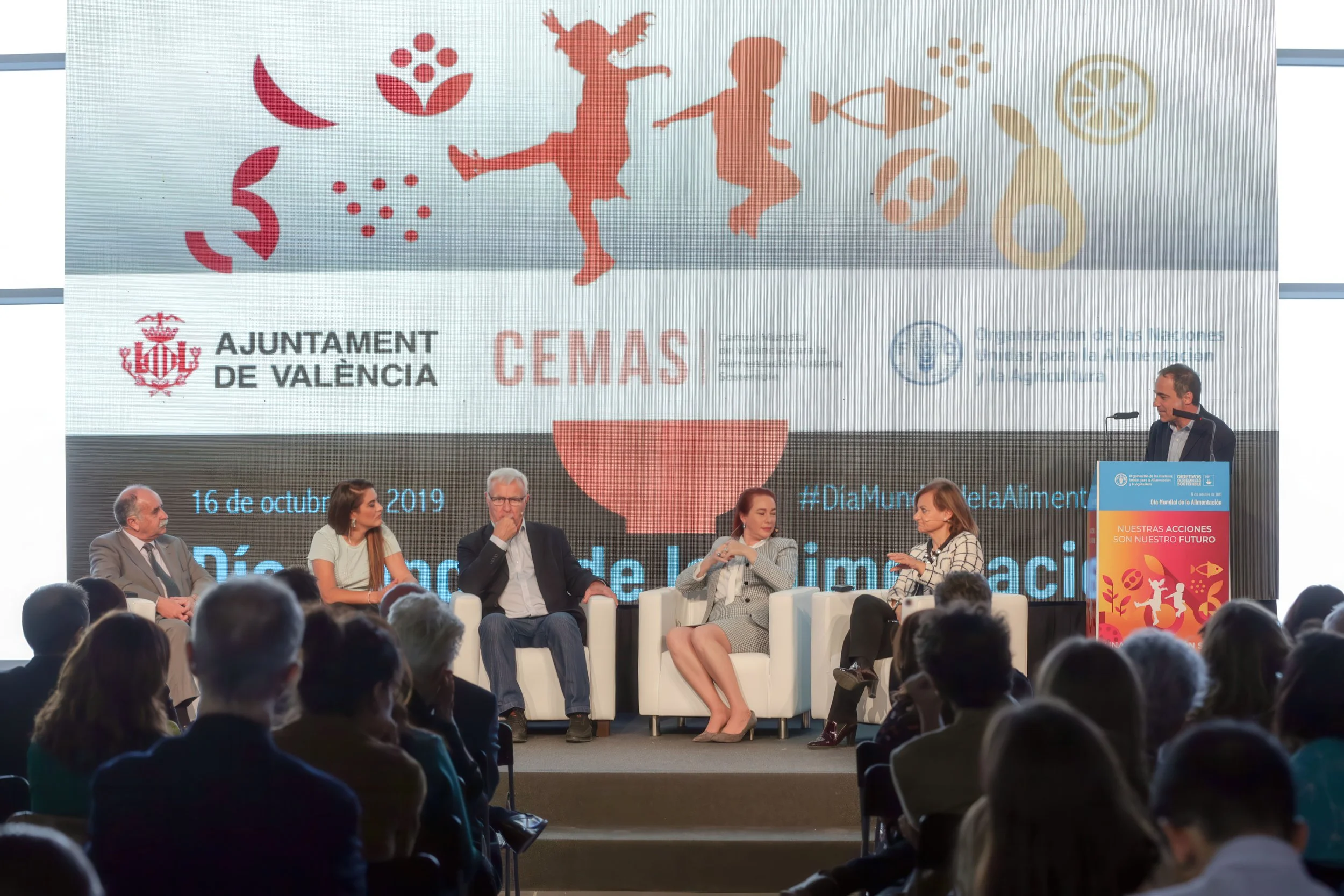 A conference or panel discussion with five people on stage, four men and one woman, seated in white chairs, with a man standing at a podium. Behind them is a large screen displaying various icons and logos related to food and nutrition, including sym