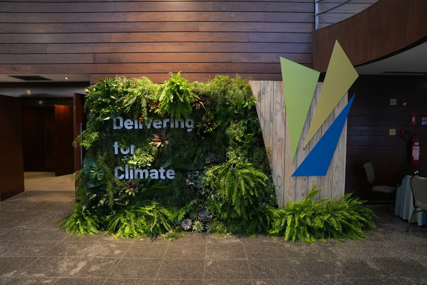 Indoor display with lush green plants and the sign 'Delivering for Climate' on a leafy wall, with a wooden and painted geometric backdrop on the right side.