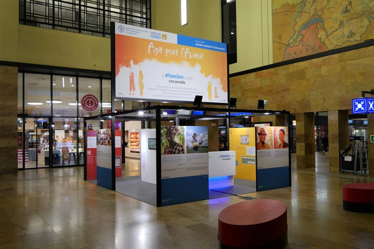 Exhibition display about the United Nations sustainable development goals in a large indoor space, featuring informational panels, photographs, and text in French, with a large screen above showing the same info.