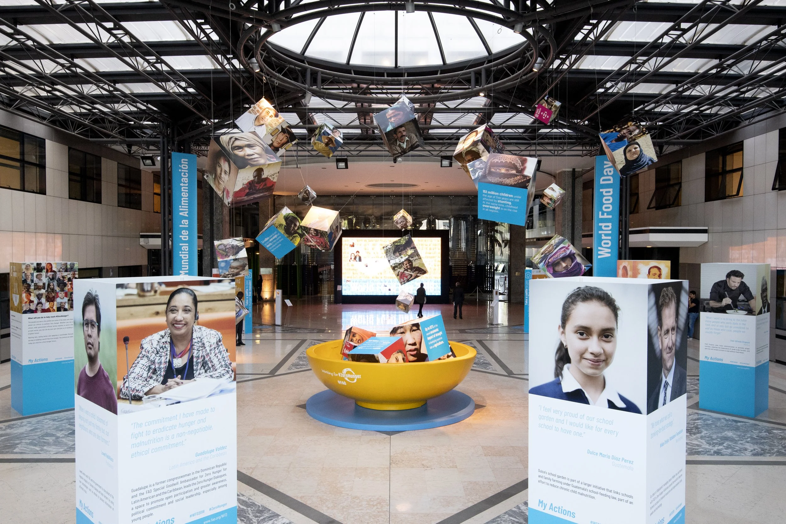 Exhibit inside a modern building features photographs of diverse children on display panels and hanging displays, with informational text about their actions and achievements, emphasizing World Food Day.