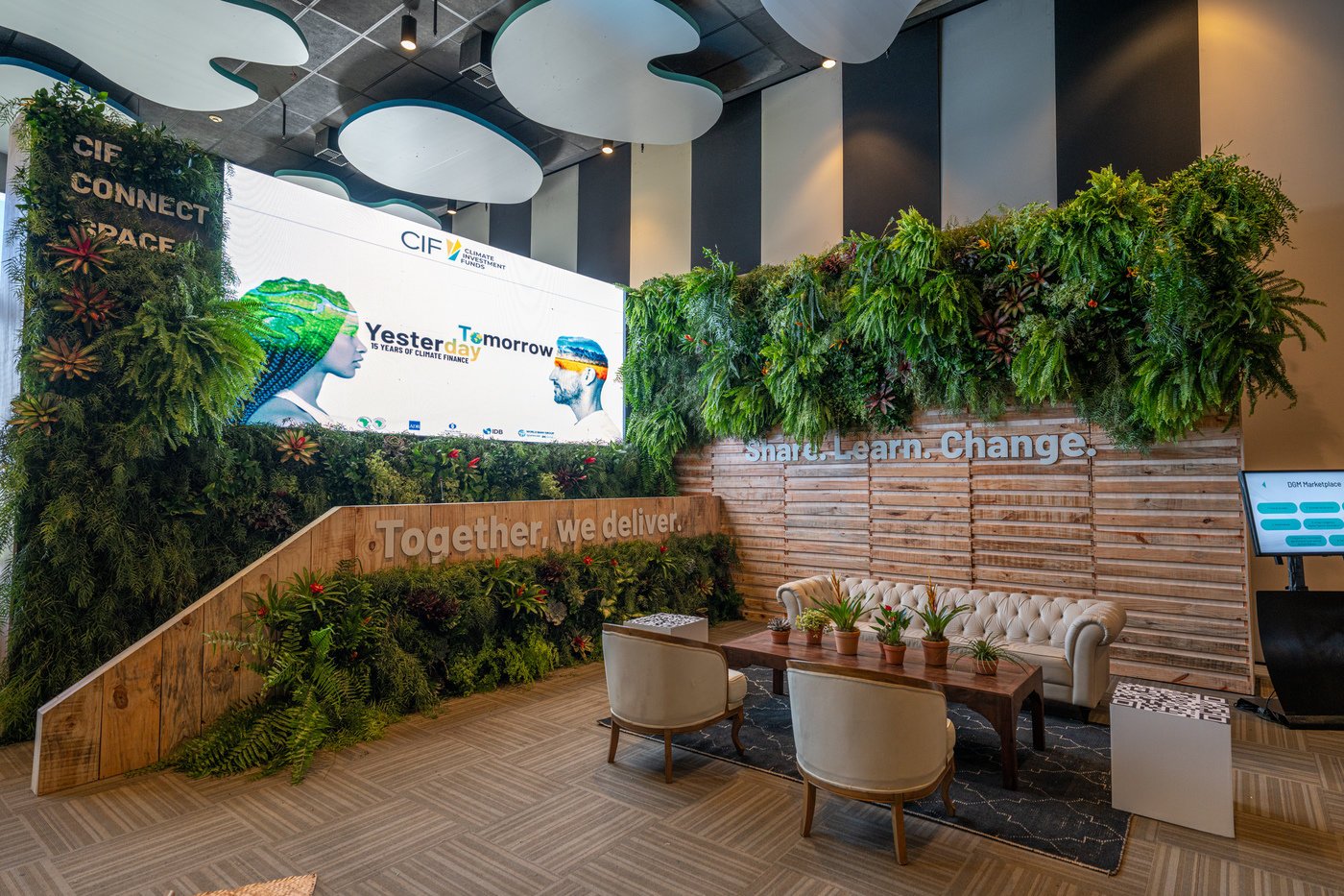 A modern indoor space with green plants and wooden walls, featuring a large screen displaying the logos of CIF and climate finance messages, along with chairs, a table with potted plants, and signs that say 'Together. we deliver.' and 'Share. Learn. 