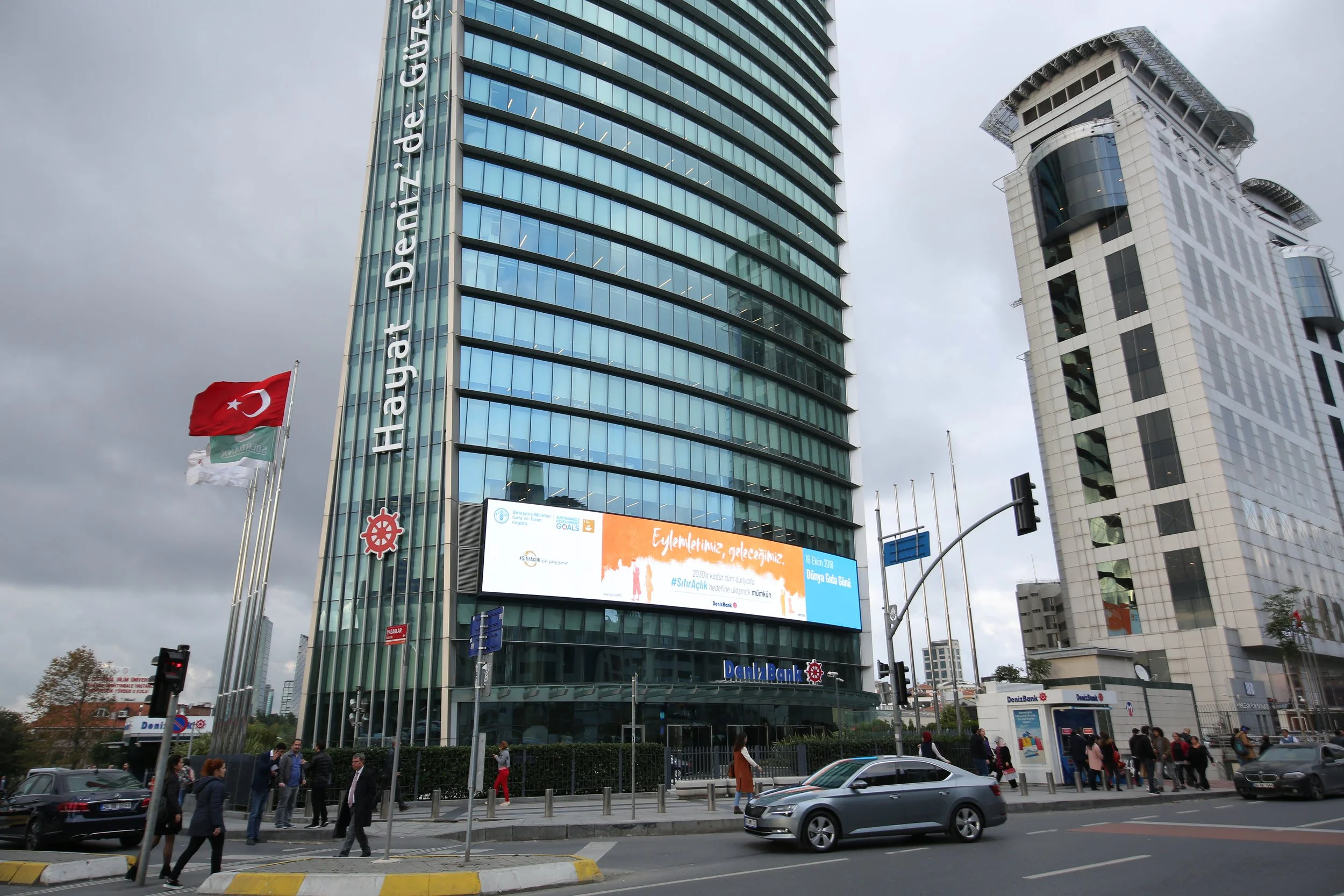 A modern glass skyscraper with the name "Dai-ichi Life GYO" on the side, in an urban setting with other buildings. Turkish flags are flying in front of the building. Several pedestrians are walking on the sidewalk, and cars are on the street. The sky