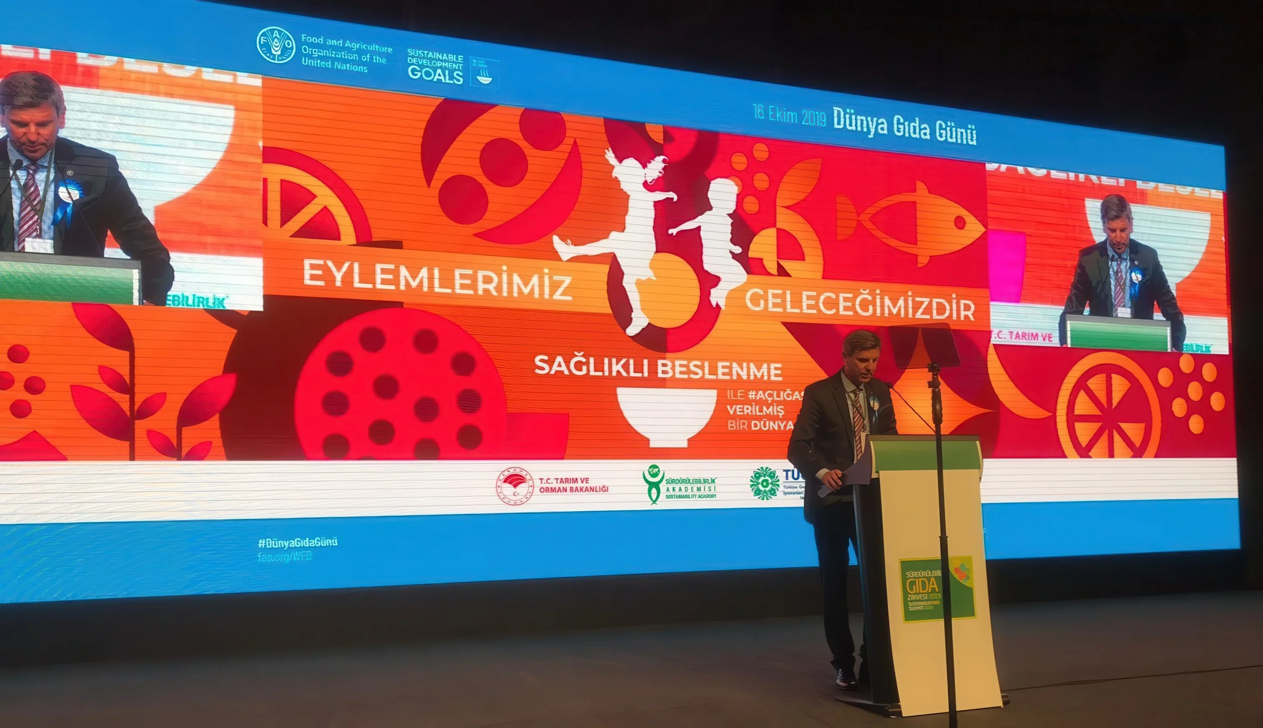 A man in a suit speaking at a podium during a conference or event related to World Food Day, with a large colorful backdrop behind him celebrating the day on October 16, 2019. The backdrop features graphics of children jumping, fish, and various abst