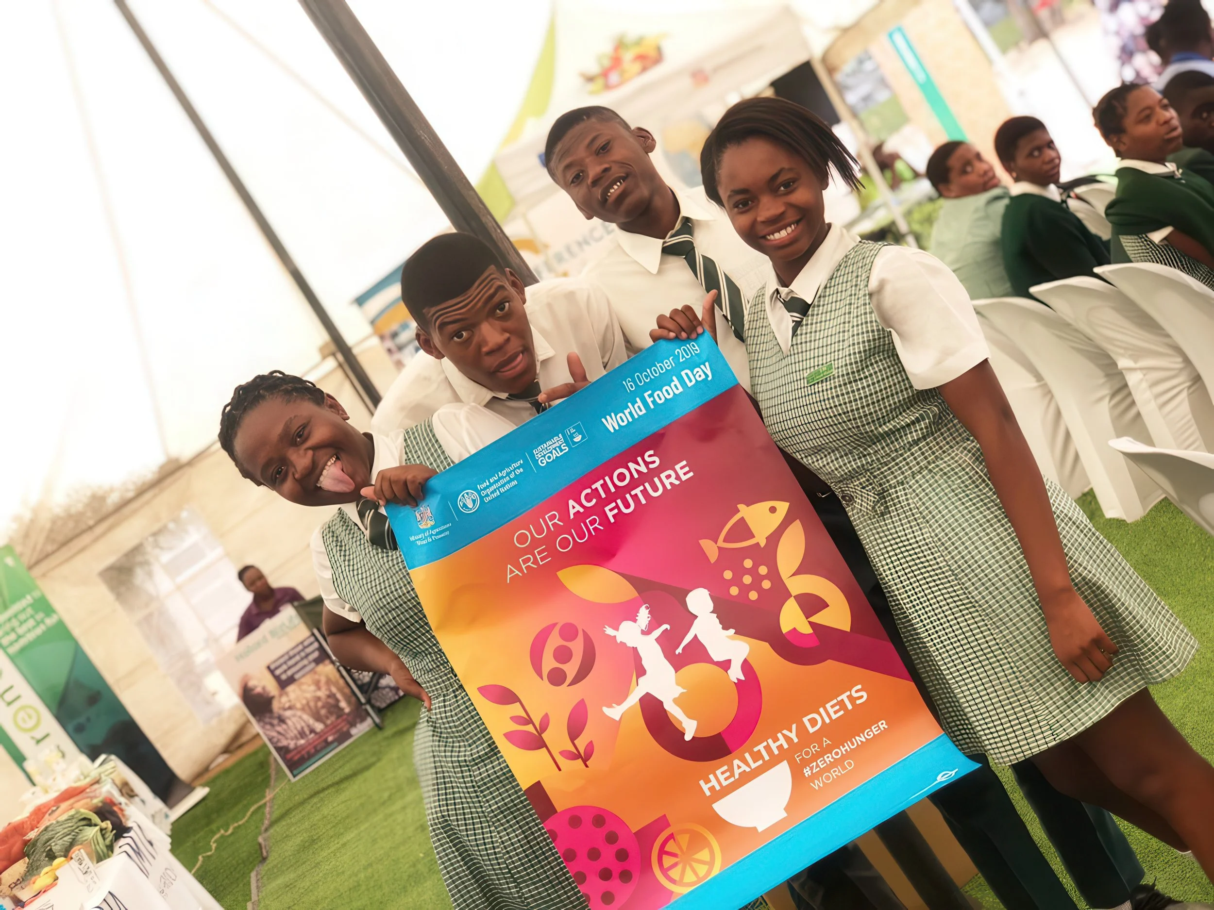 School children at a booth for World Food Day event holding a colorful poster promoting healthy diets and zero hunger, with other booths visible in the background.