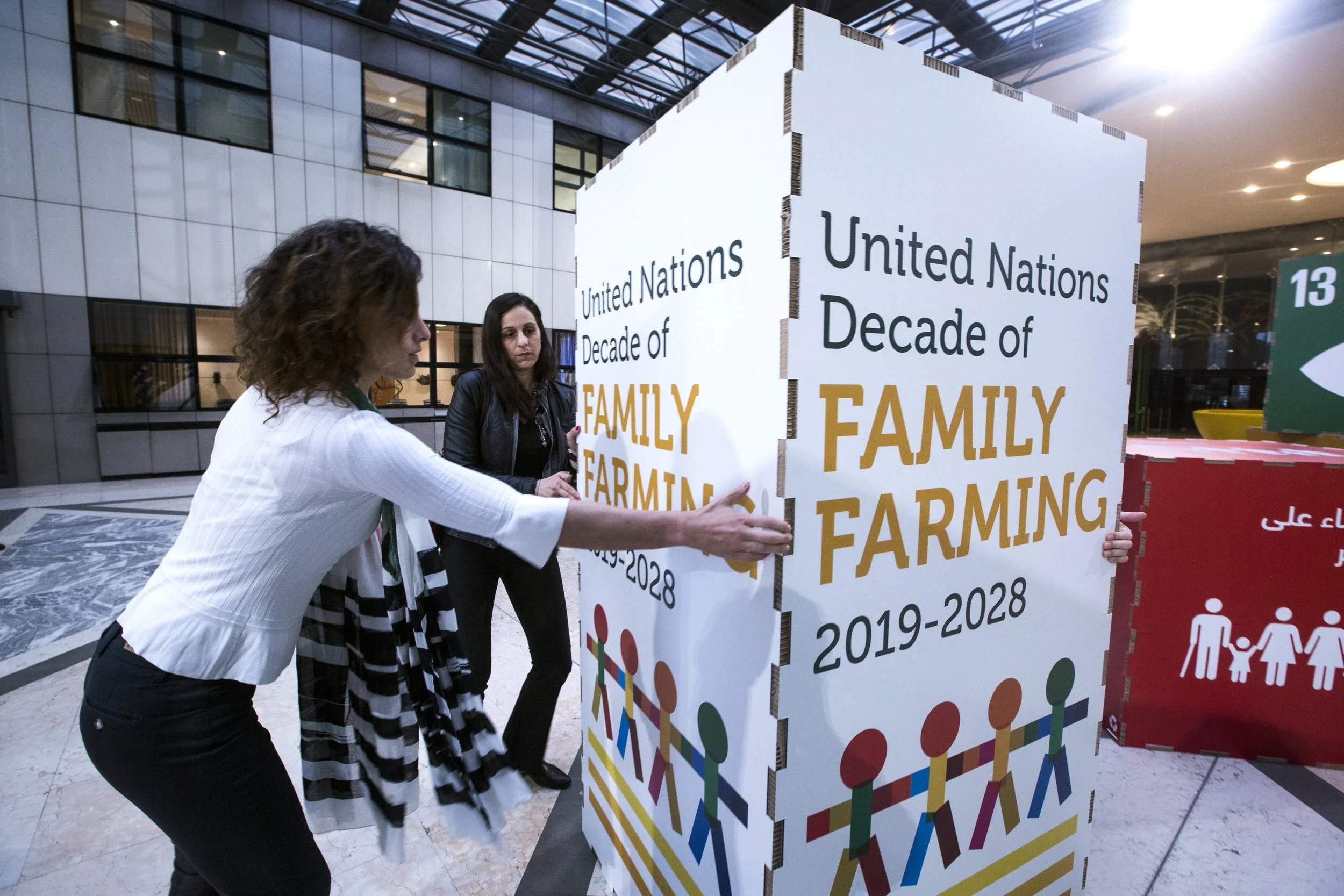 Two women placing a sign at the United Nations marked with text indicating a decade of family farming from 2019 to 2028, inside a modern building with glass windows and a high ceiling.