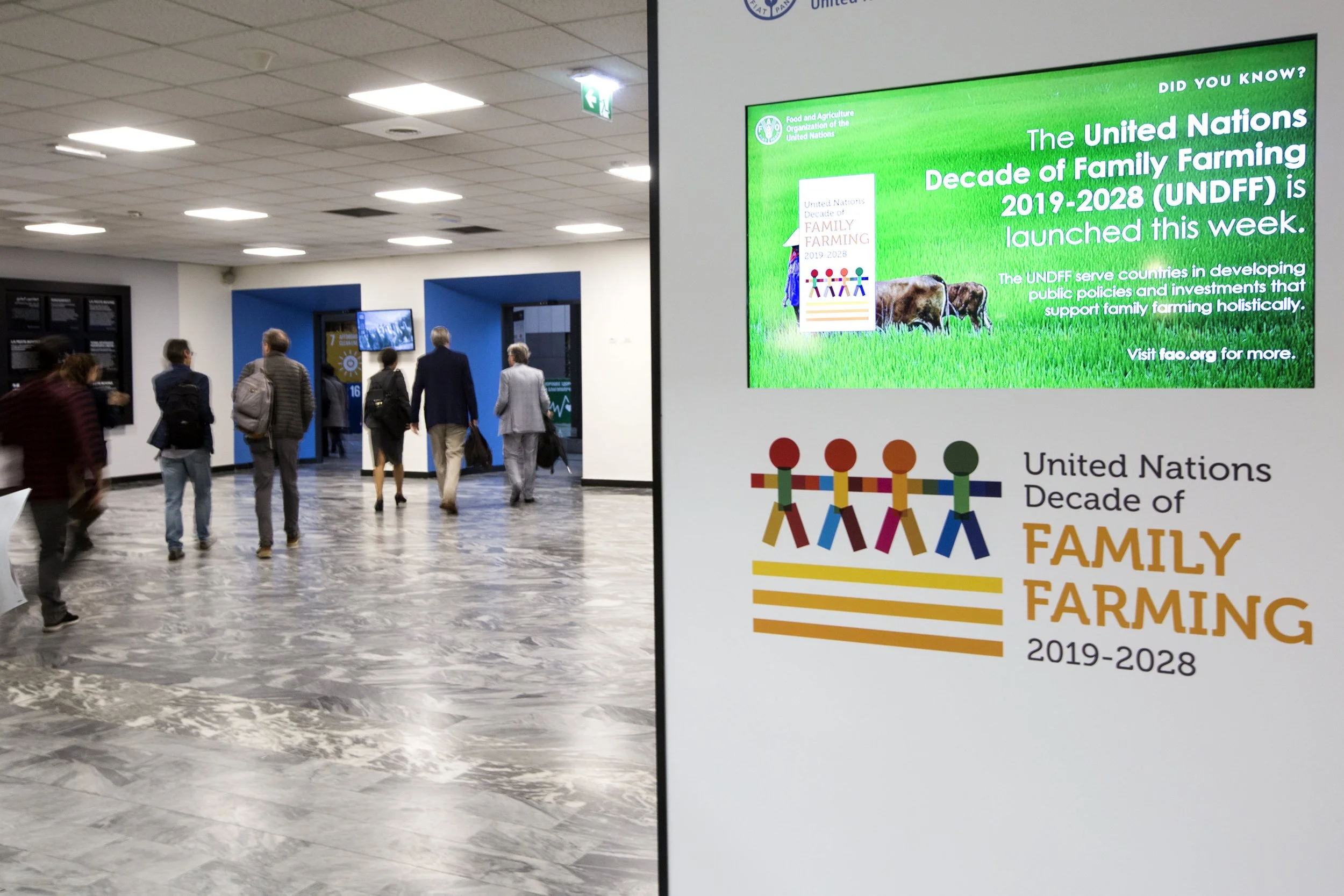 An indoor hallway with people walking, next to a digital display promoting the United Nations Decade of Family Farming 2019-2028 initiative.