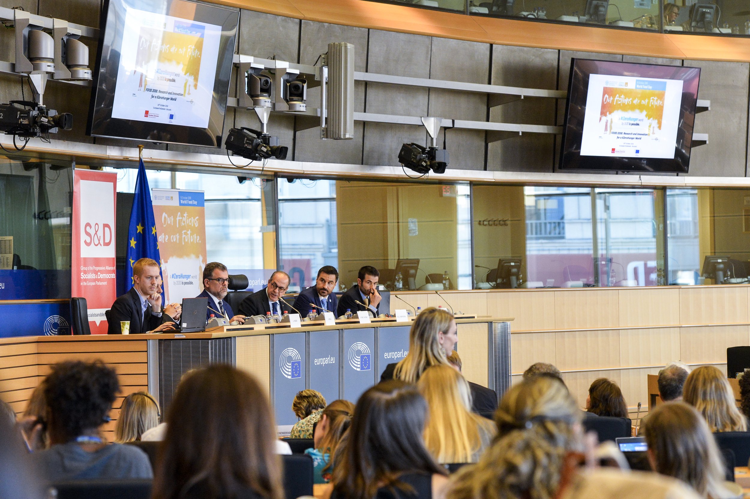 A panel of five people sitting at a desk during a conference or meeting at the European Parliament. The background displays banners and screens with text about food research and innovation, and the European Union flag is visible.