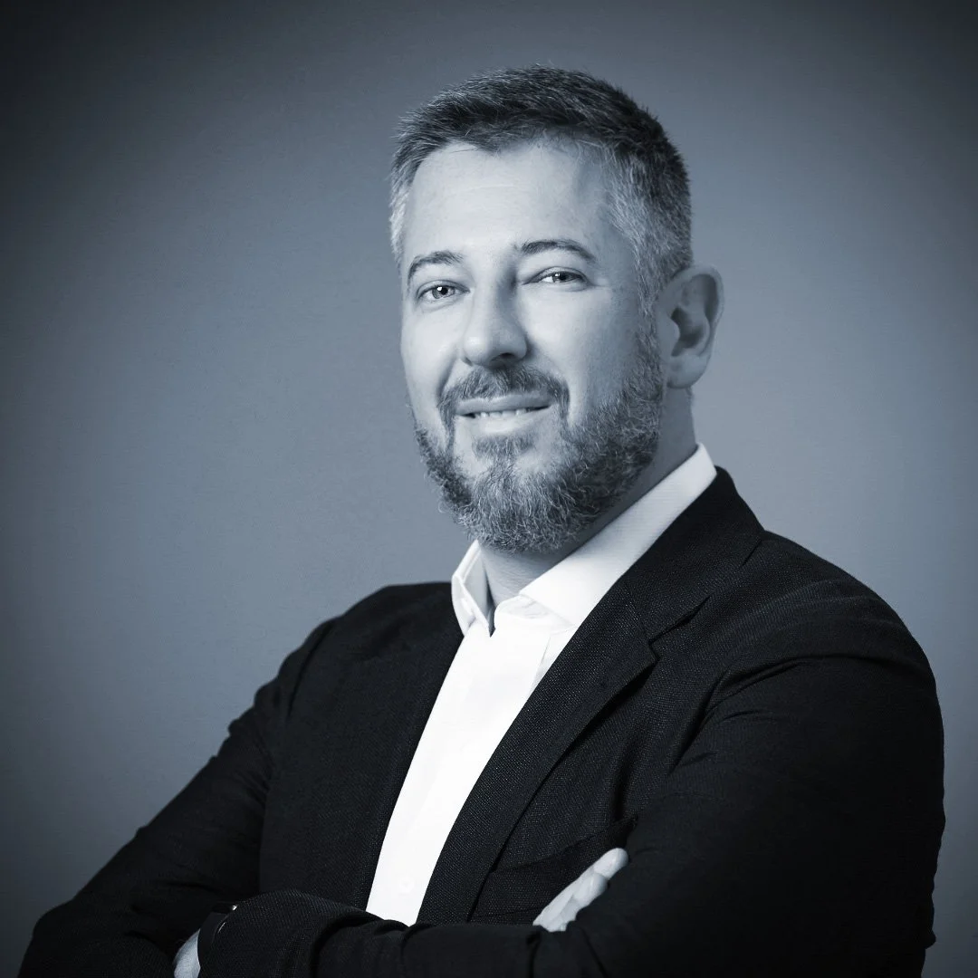 Black and white professional headshot of a man with short hair and a beard, wearing a dark blazer and white shirt, smiling with arms crossed.