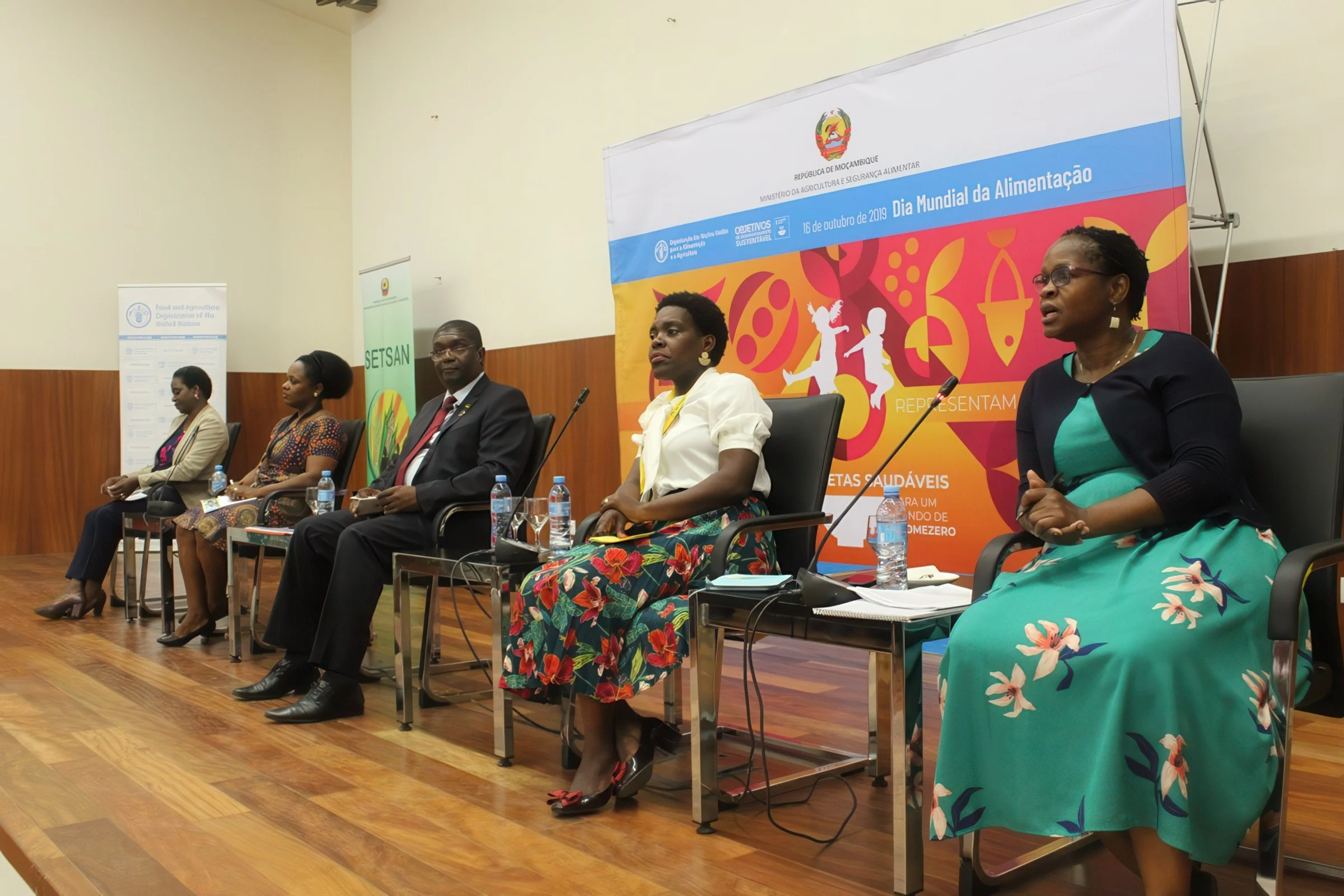 A panel of five people sitting on a stage at a conference, with a colorful banner behind them. The banner displays the logo of the Republic of Mozambique and mentions World Food Day on October 16, 2019. The panel includes four women and one man dress
