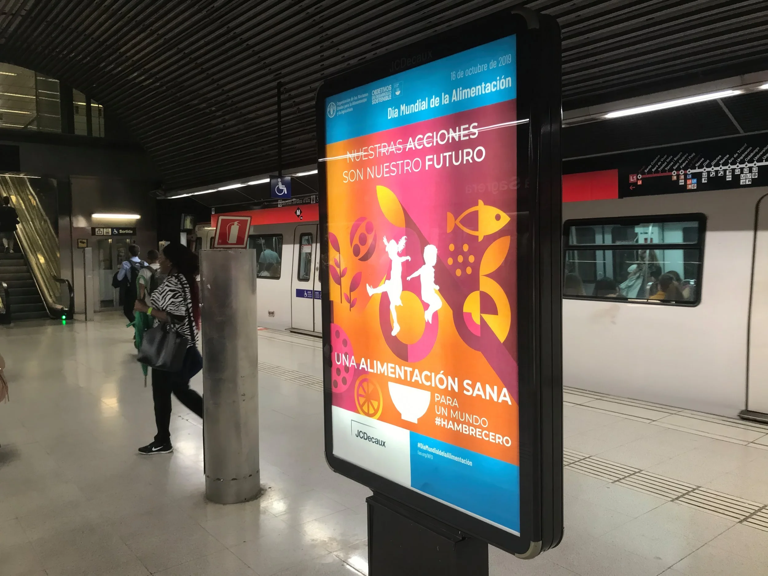 Digital advertisement poster at a subway station for World Food Day, promoting healthy eating habits for a sustainable future, with illustrations of a fish, a bowl of food, and children playing.