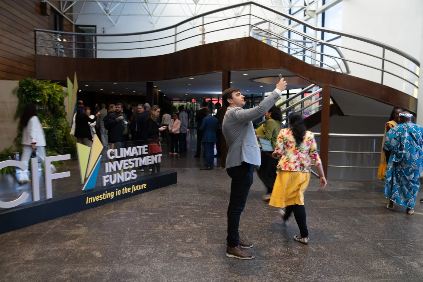At a conference, a group of people gathers in a modern, indoor venue with a staircase. A prominent sign reads 'Climate Investment Funds' with a tagline 'Investing in the future.' One man in a gray blazer is taking a selfie or photo, while others are 