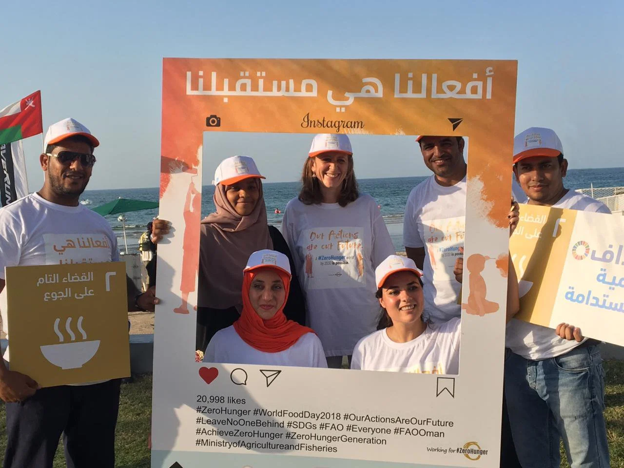 Group of six people at the beach holding a large Instagram-style photo frame with Arabic and English text for World Food Day 2018, promoting zero hunger.