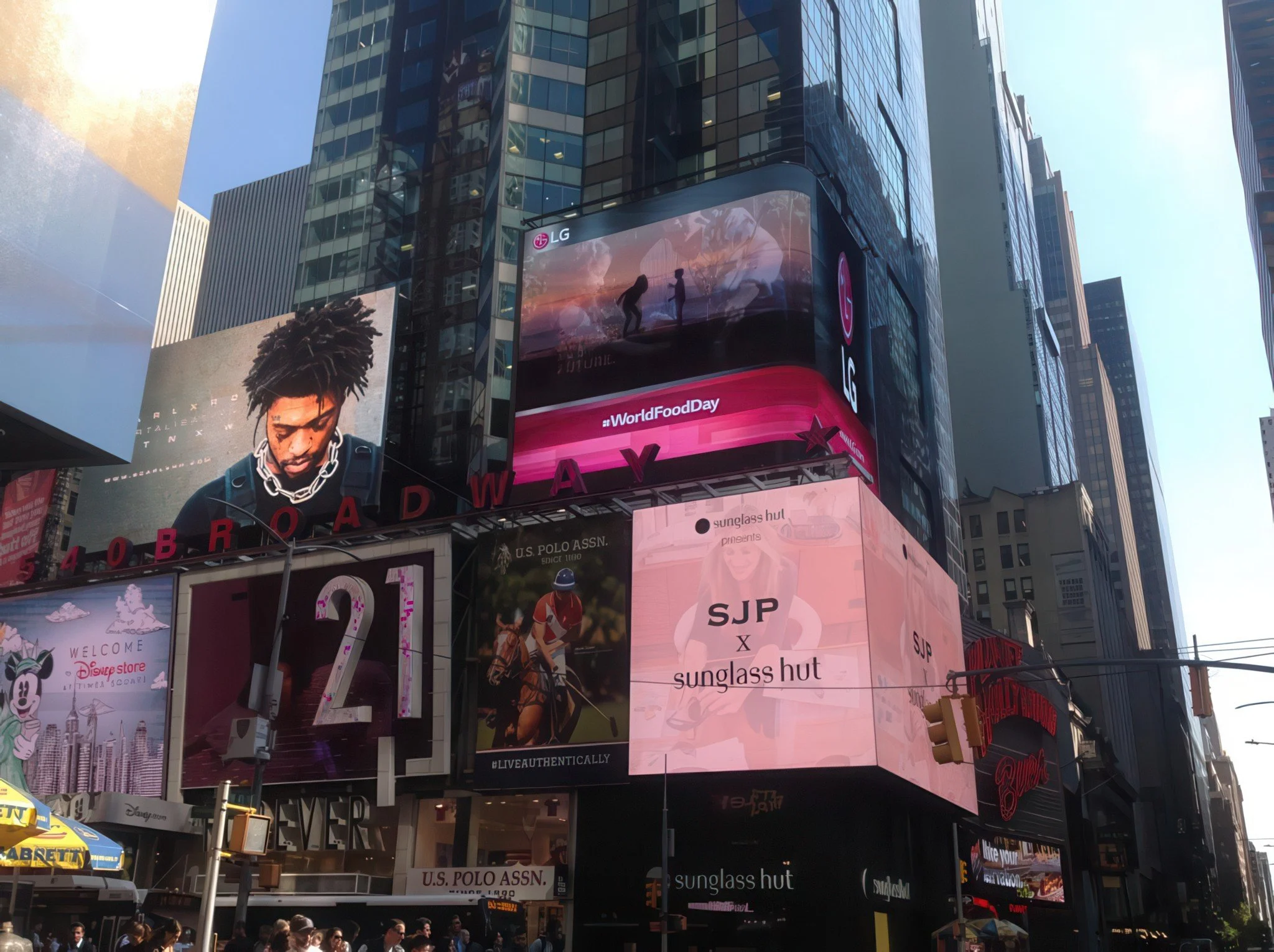 Tall building with multiple digital billboards displaying advertisements, including one for LG, one for Sunglass Hut, and a music video with two silhouette dancers. Crowds of people at street level.
