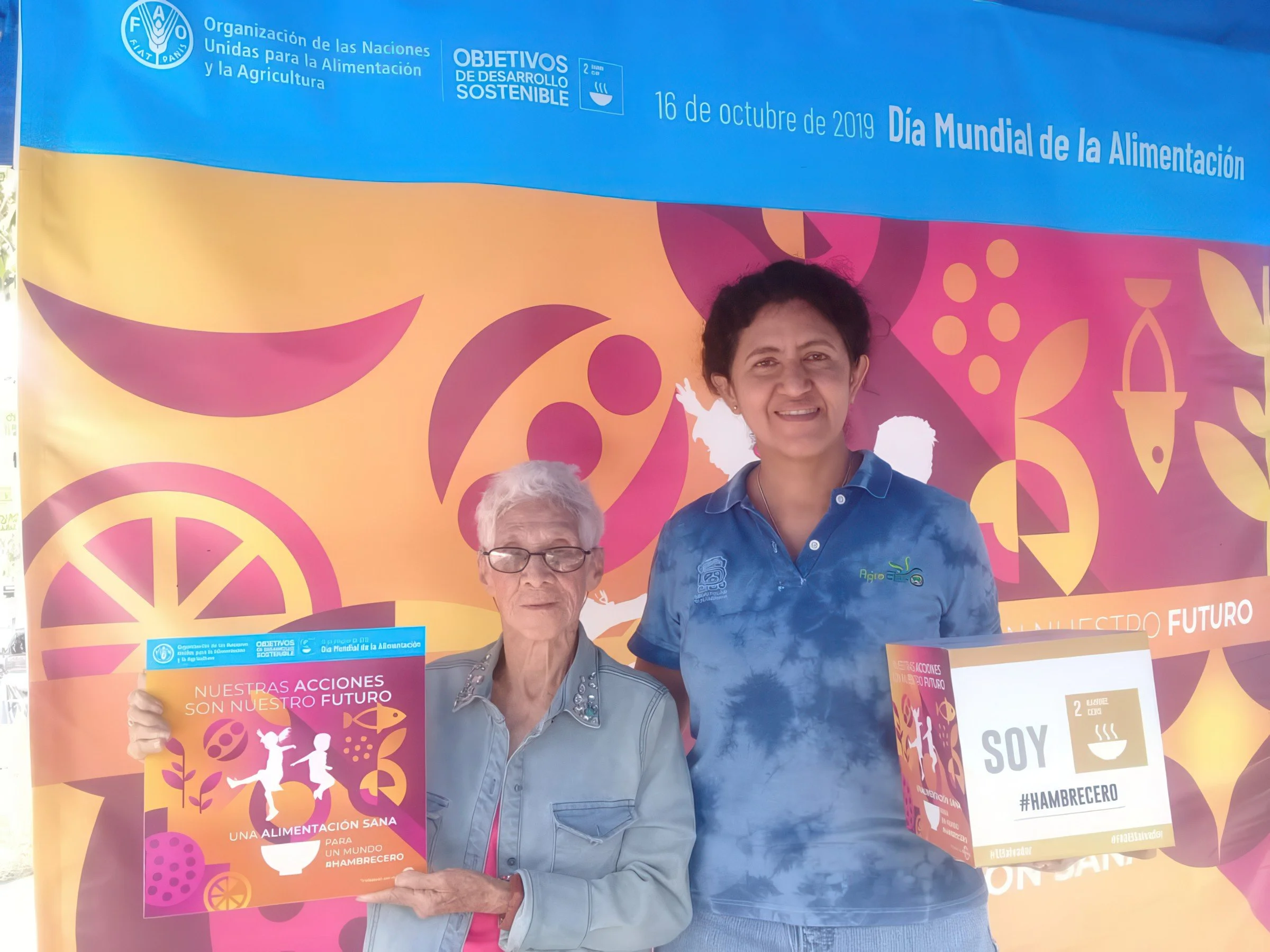 A woman and an older woman holding a sign in front of a colorful background with signs about World Food Day, sustainable development goals, and healthy eating.