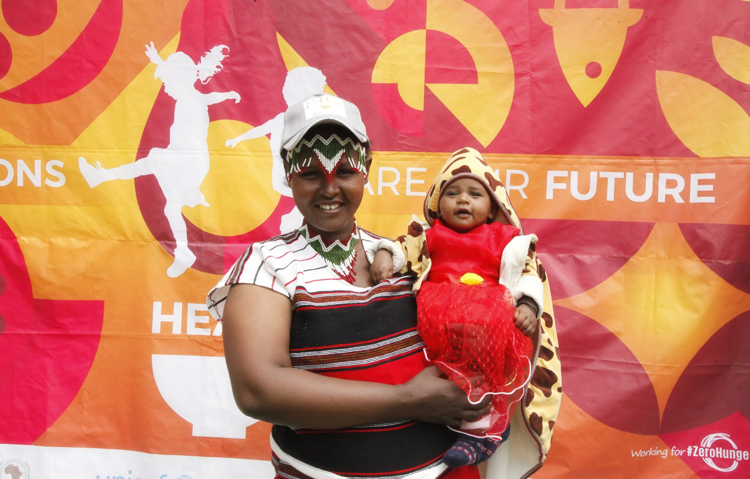 A woman holding a child in front of a vibrant red, orange, and yellow backdrop with abstract shapes and a white silhouette of a dancer. The woman and child are wearing animal print and striped clothing. The backdrop features the text 'Working for #Ze