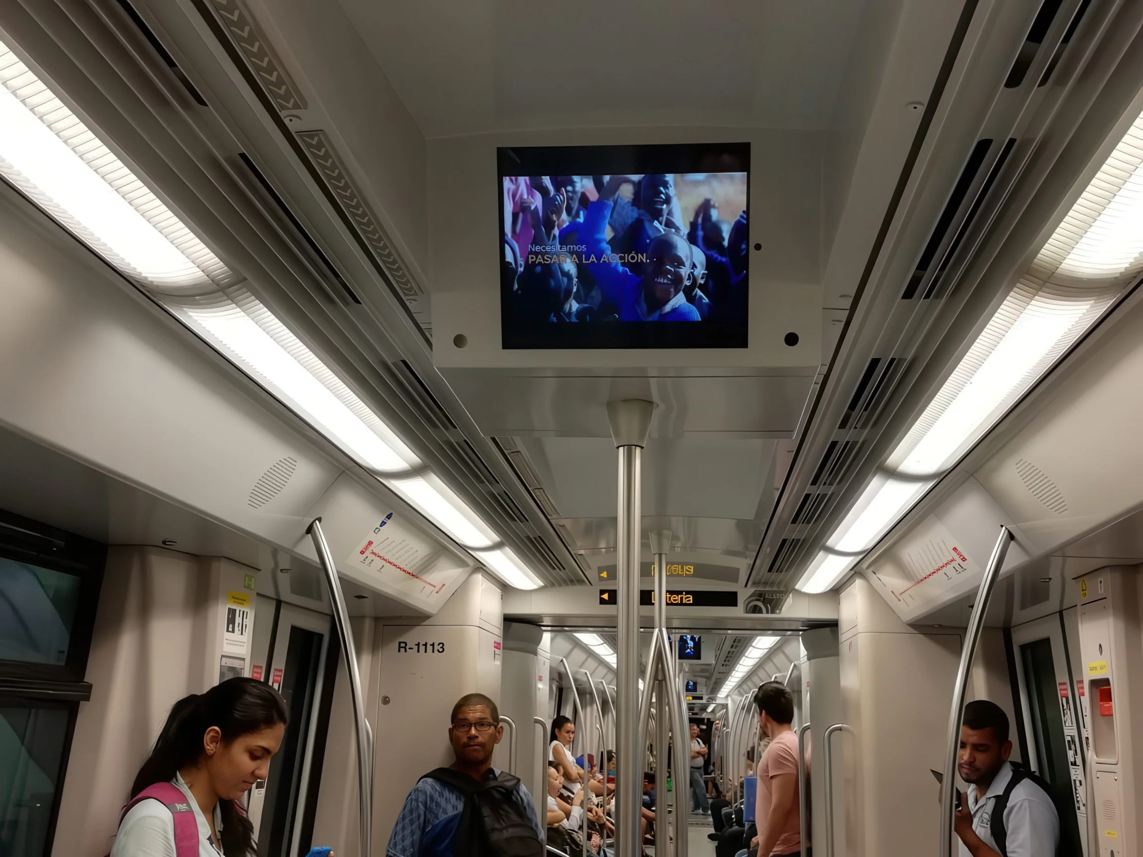 People standing and sitting inside a modern subway train with overhead digital display showing an advertisement and a screen displaying a crowd of smiling children.