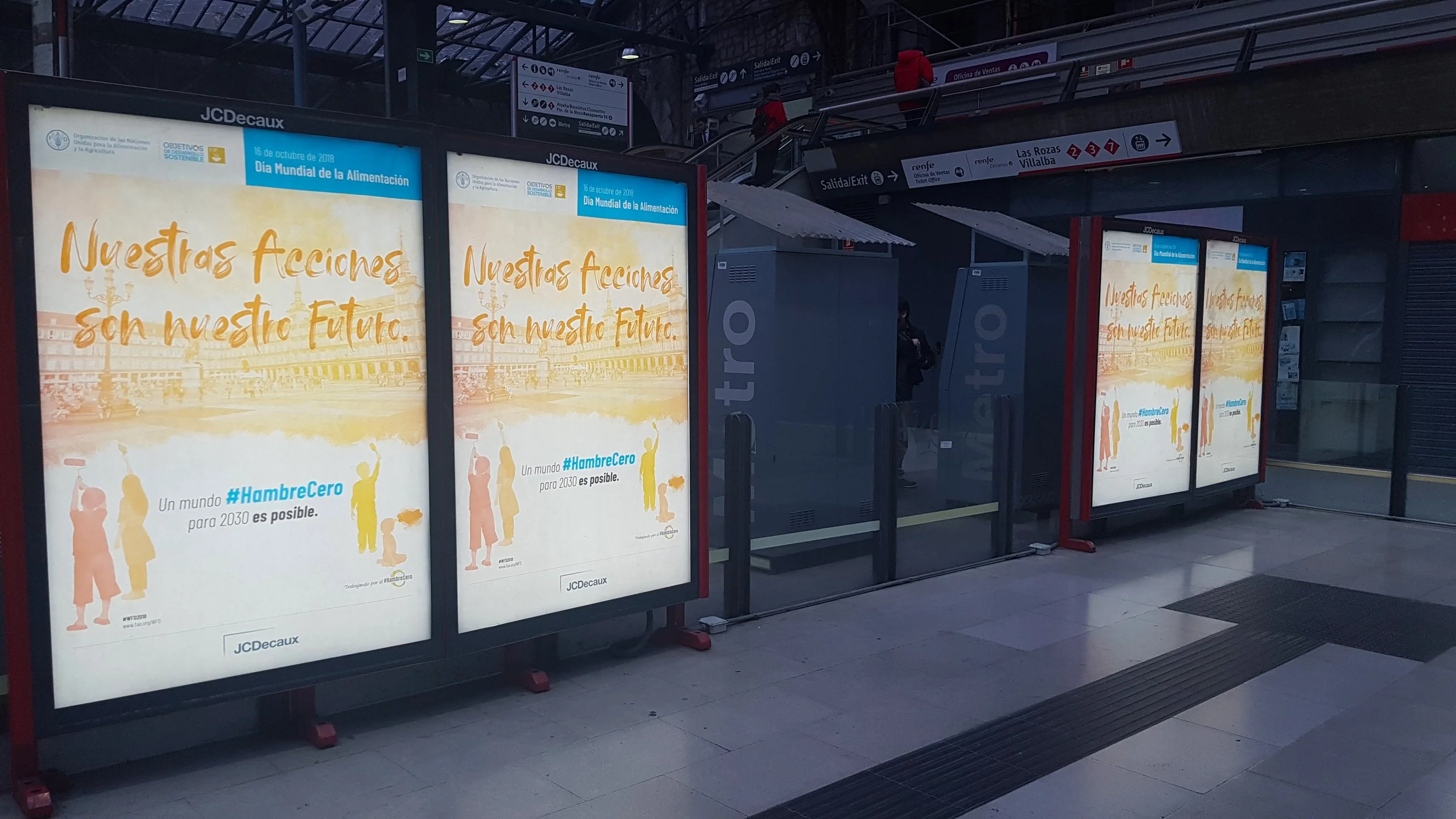 Bus stop with digital billboards displaying an advertisement in Spanish for World Food Day, featuring illustrations of people and the hashtag #HambreCero.