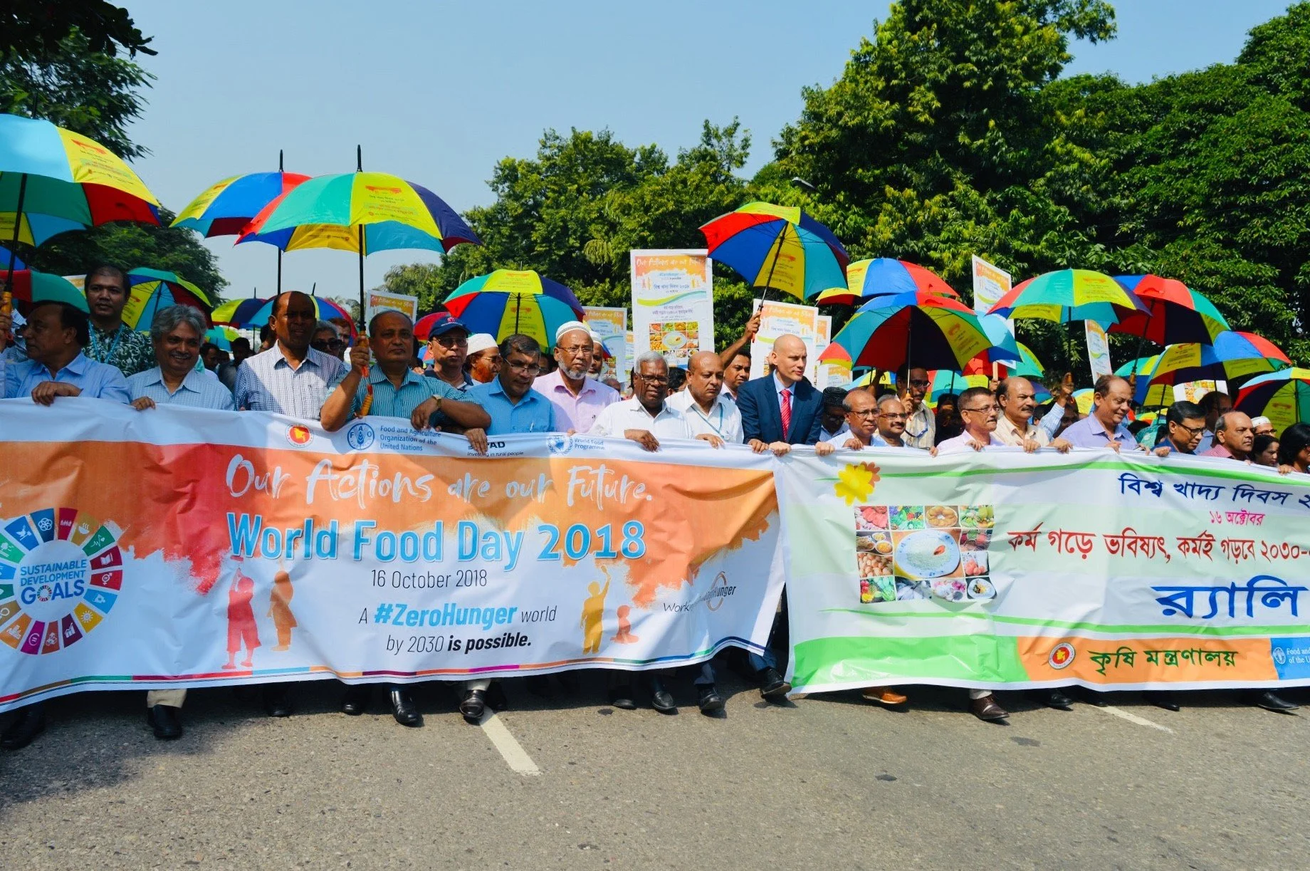 A group of people participating in a parade or demonstration holding banners and colorful umbrellas during a celebration for World Food Day 2018 on October 16, 2018, outdoors on a sunny day with green trees in the background.