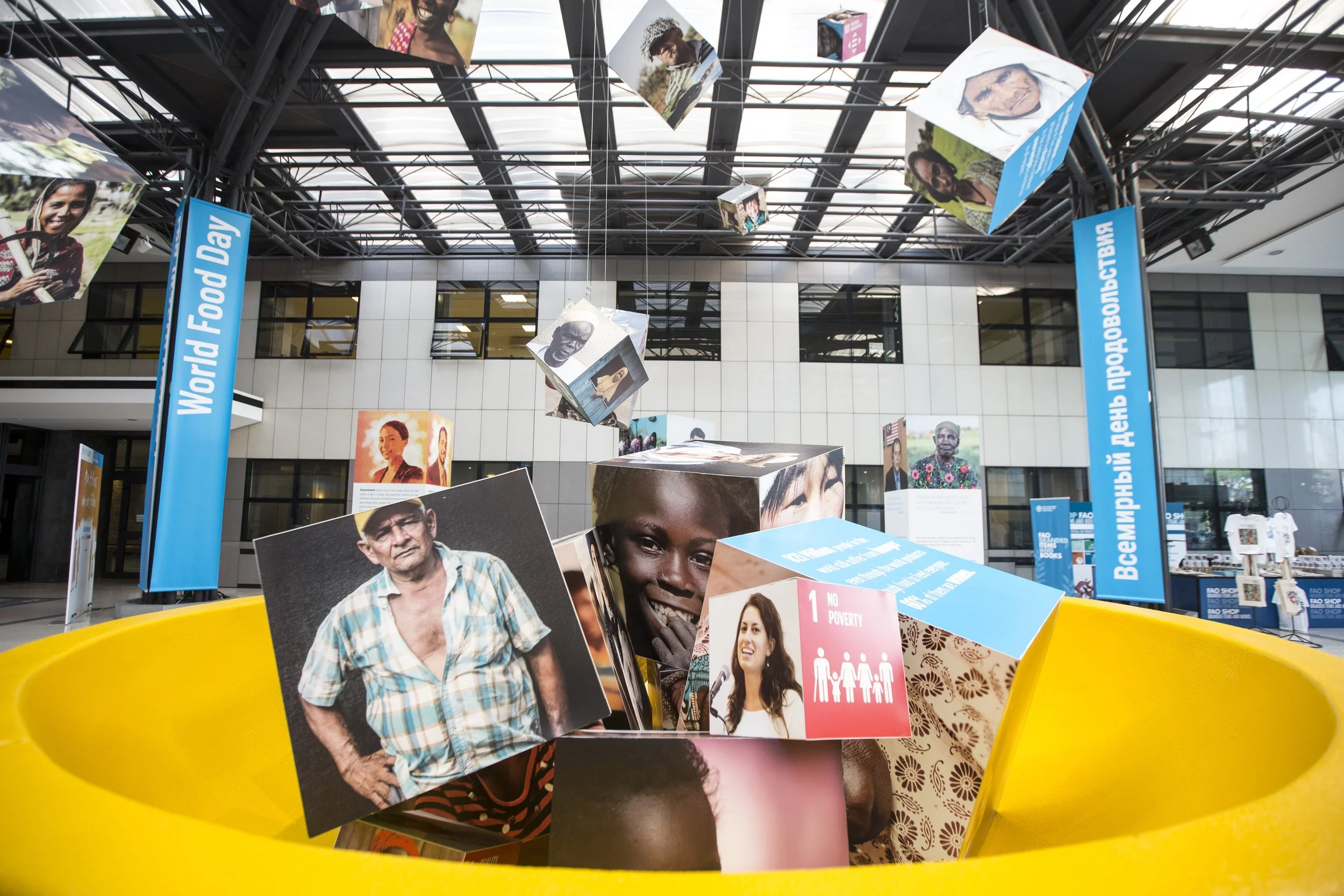 Exhibit display with hanging photographs of diverse individuals inside a modern building, with blue informational banners on either side and a yellow circular barrier at the bottom.