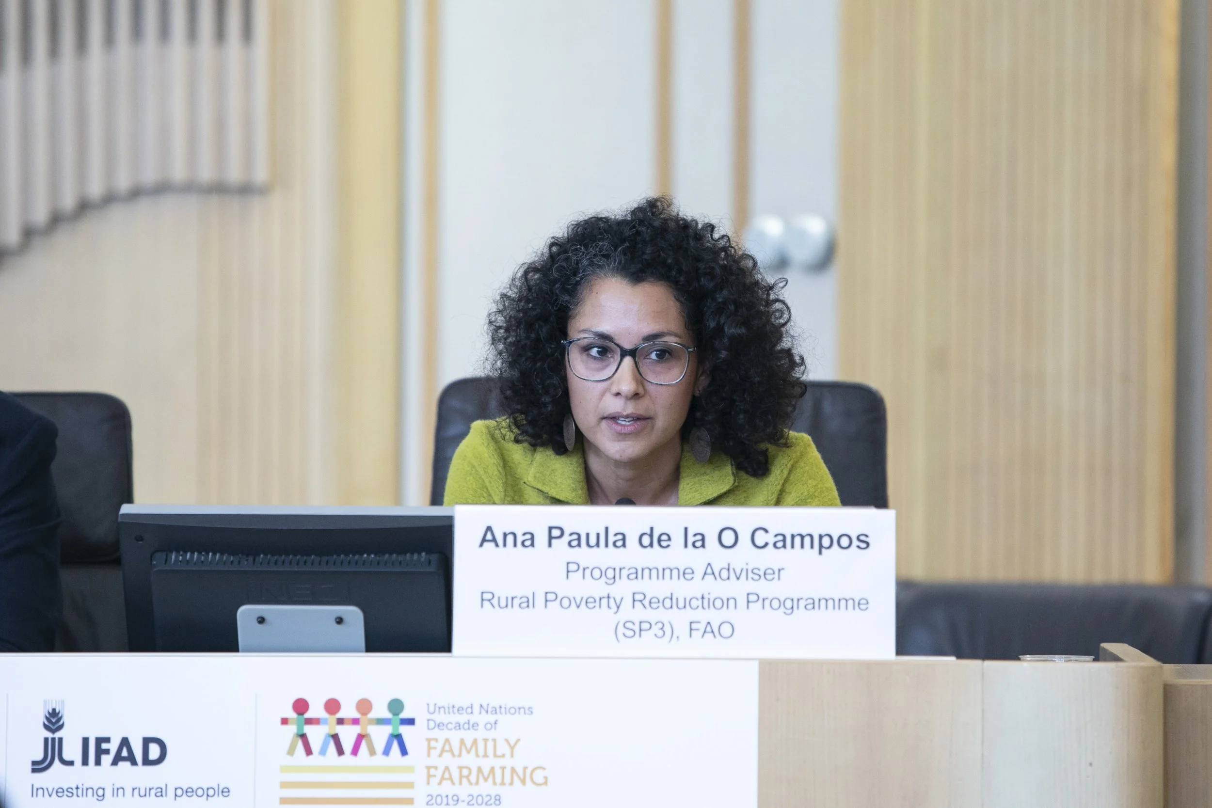 A woman with curly dark hair and glasses wearing a green jacket, sitting at a desk with a sign that reads 'Ana Paula de la O Campos, Programme Adviser, Rural Poverty Reduction Programme, FAO.'