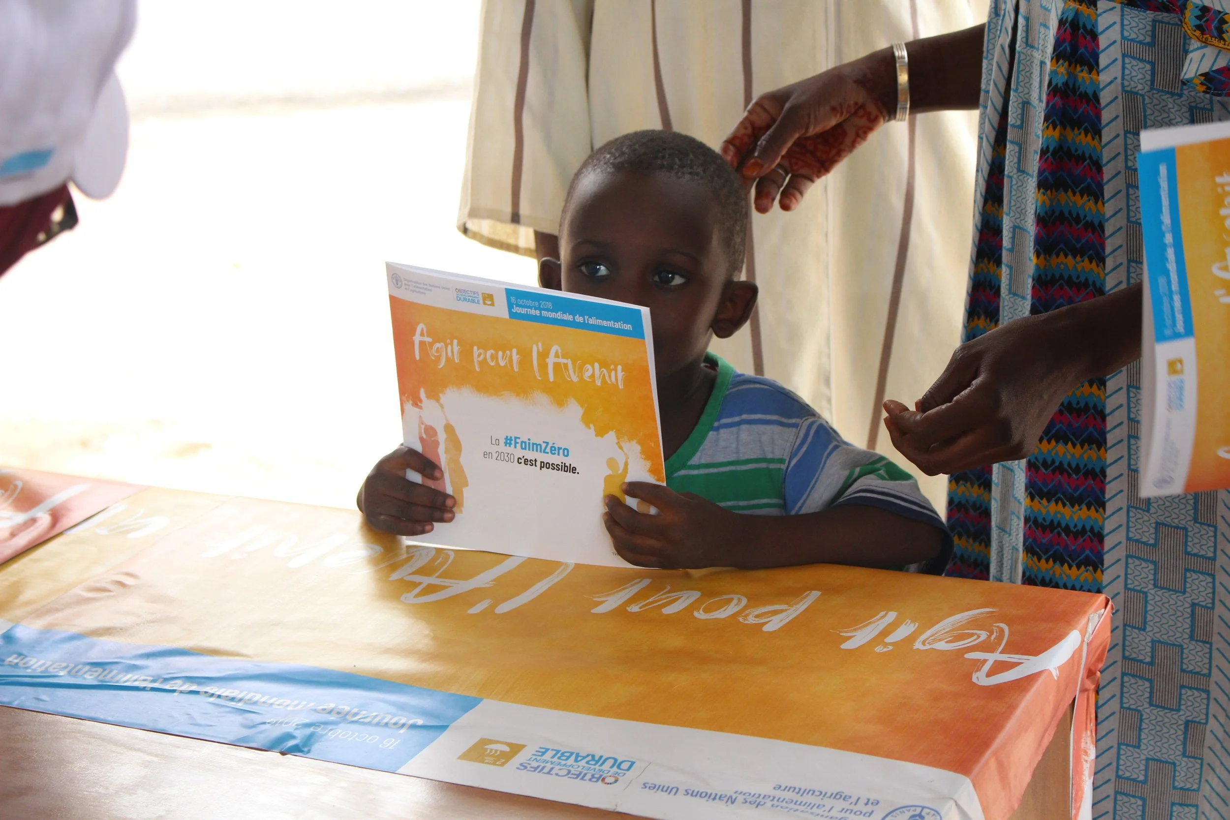 A young boy in a striped shirt is sitting at a table holding a booklet about sustainable development goals, with an adult woman wearing a colorful patterned dress standing beside him, touching his head gently.