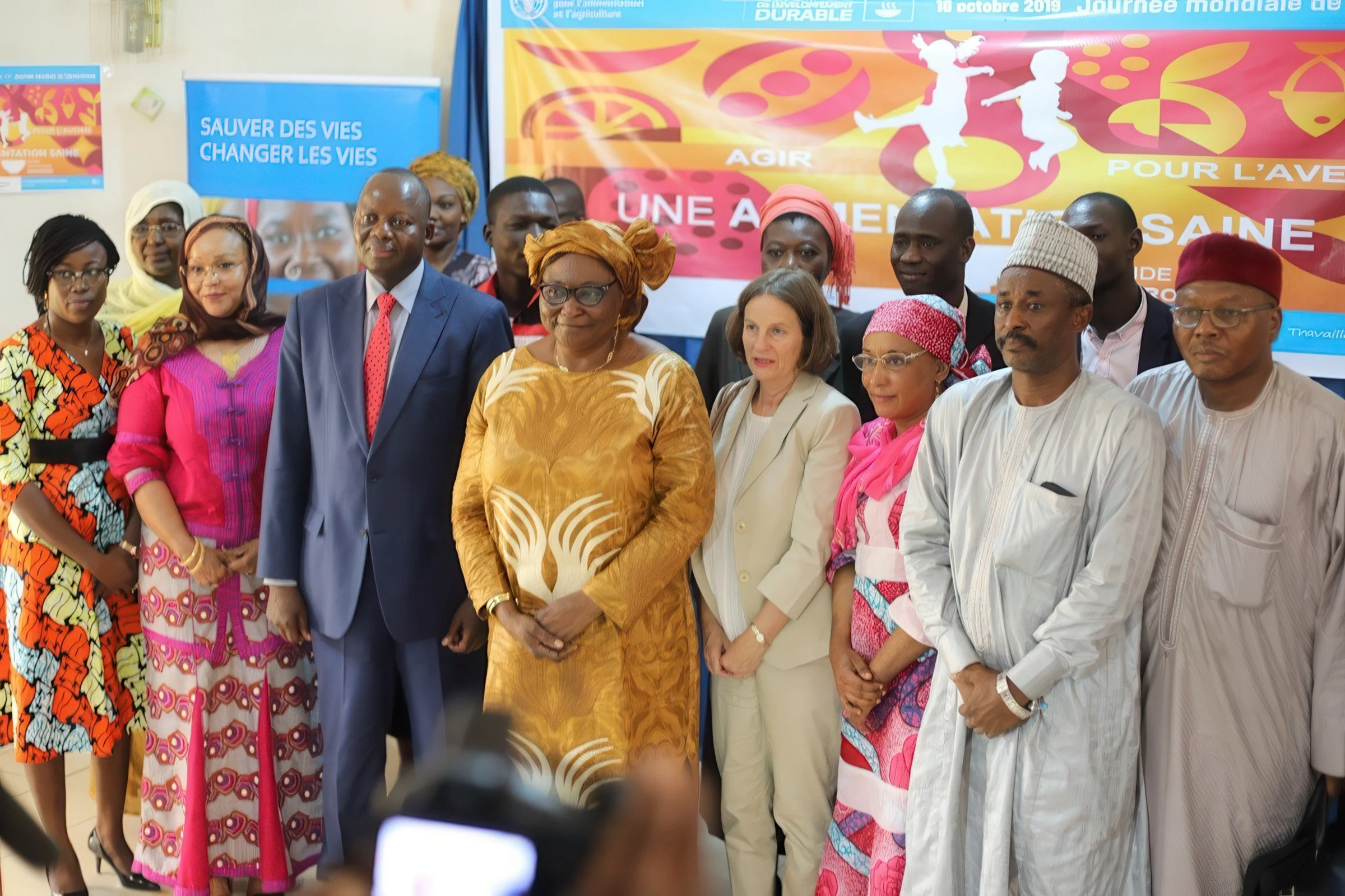 Group of people dressed in colorful formal attire standing together at an event, with banners in the background promoting health and life-saving messages.