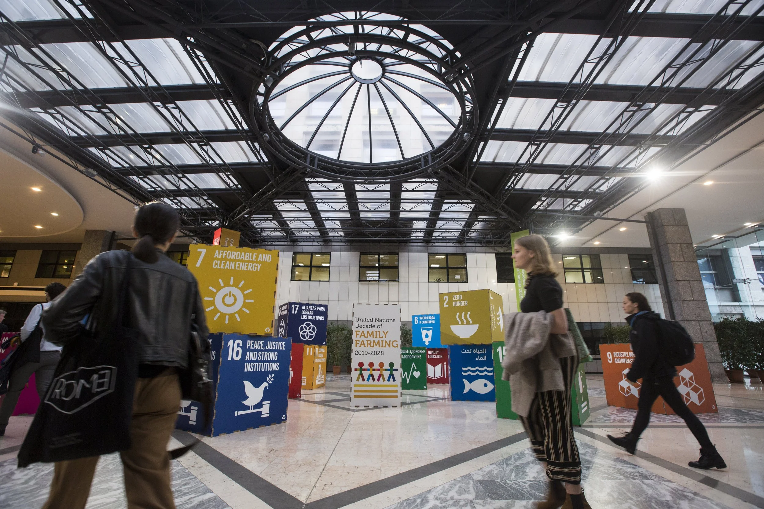 People walking in a modern indoor space with various colorful signs about social and environmental issues, above a glass and metal ceiling structure.