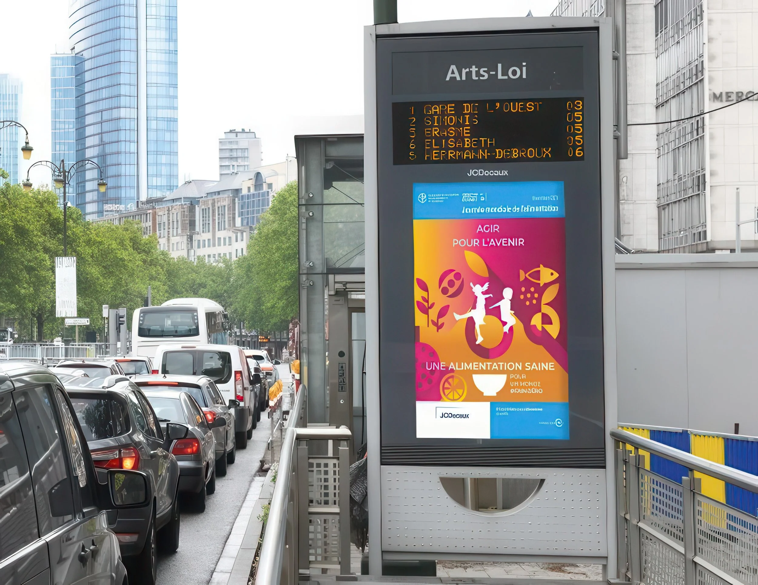 City street scene showing parked cars along a sidewalk, a digital bus stop display, and tall modern buildings in the background.