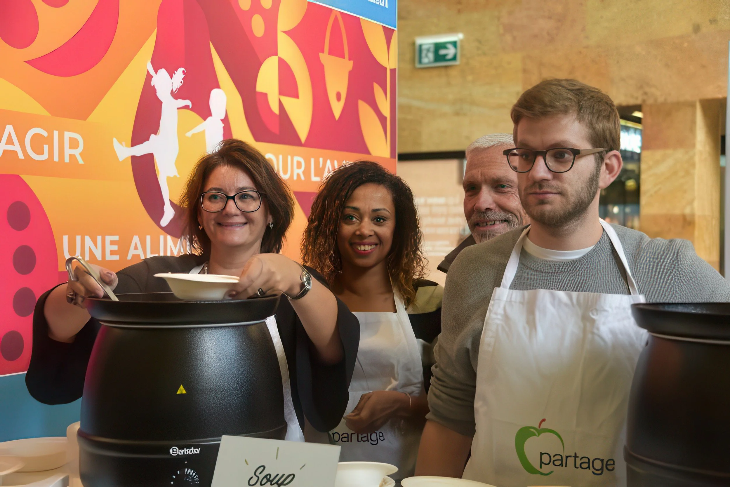 Group of people at a food event, pouring soup from a black pot, with colorful signage in the background.