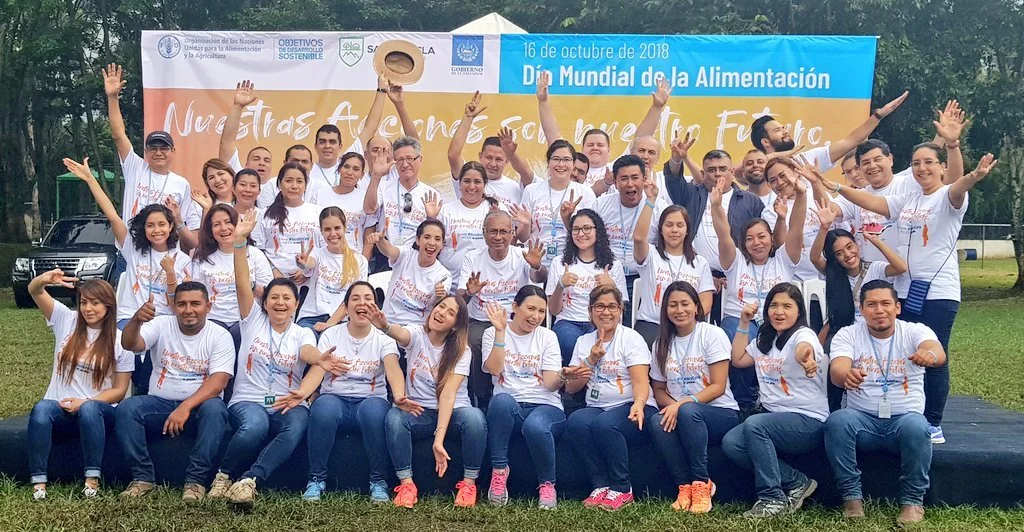 Group of people wearing white shirts posing outdoors for a photo during an event, with a banner in the background that reads '16 de octubre de 2018 Día Mundial de la Alimentación' and other Spanish text.