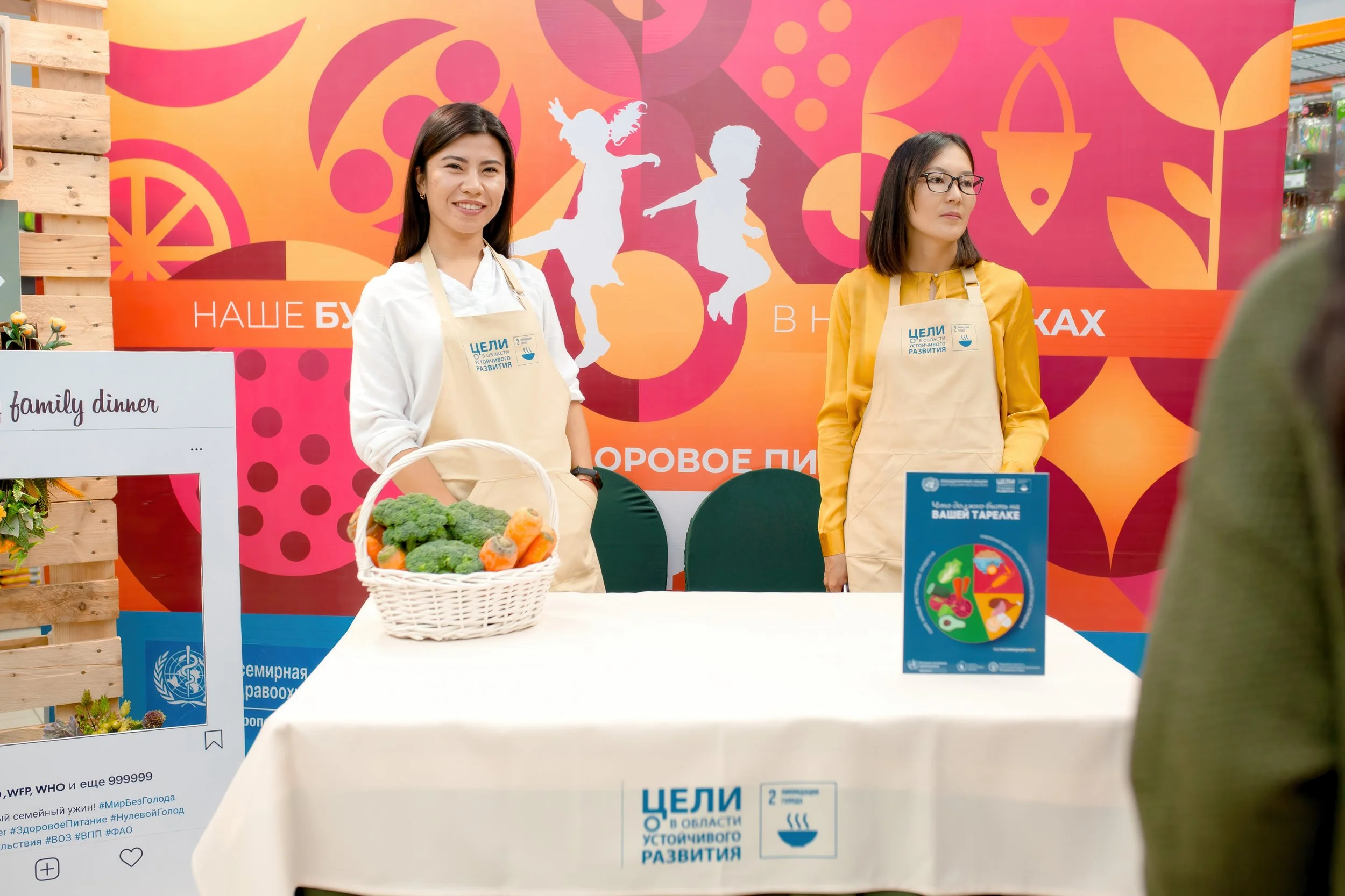Two women standing behind a table at a booth, with a basket of fresh vegetables including broccoli and carrots, and a colorful backdrop with abstract shapes and Russian text promoting sustainable development and healthy eating.
