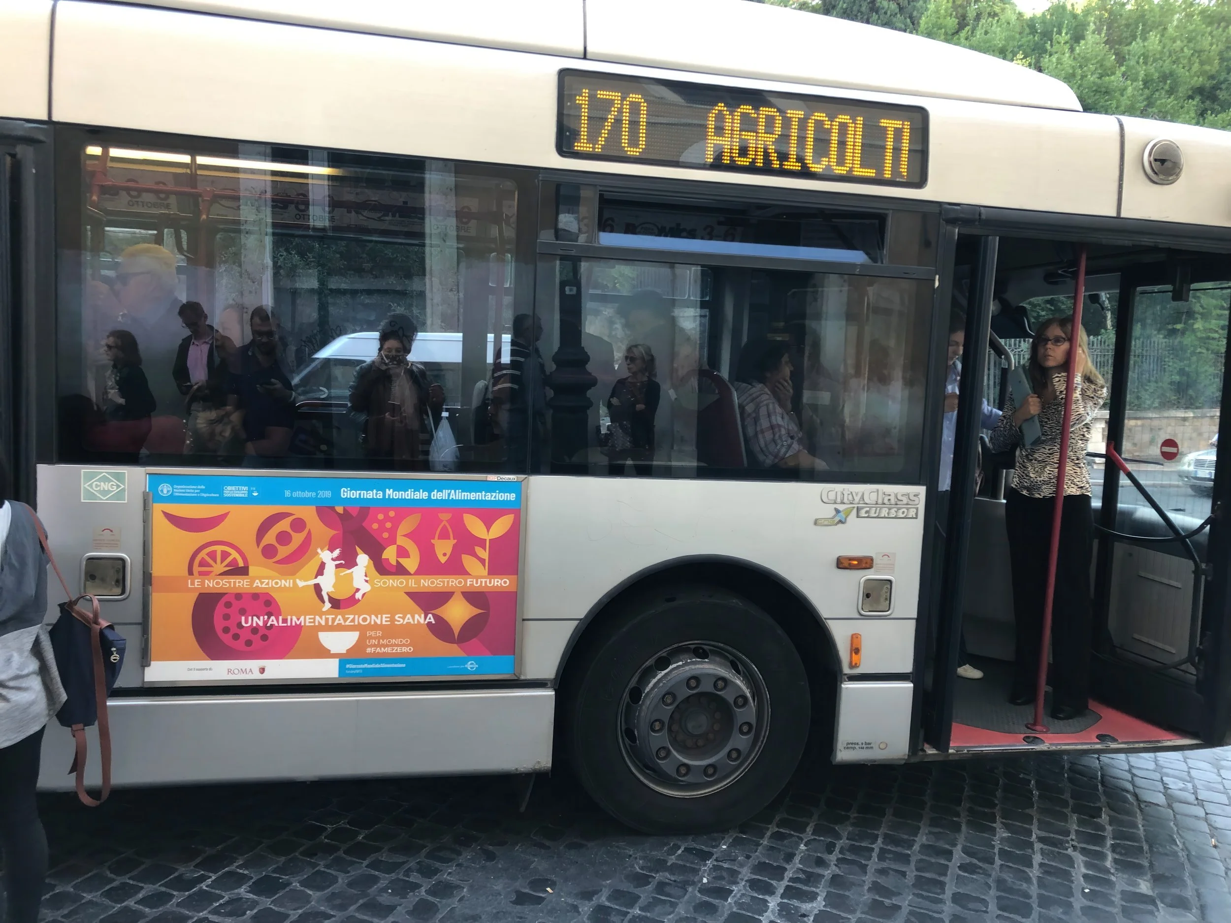 A city bus with passengers inside, some standing and some seated, waiting at a bus stop. The bus displays the route number 170 and the destination 'Agrisolt'. There is a colorful advertisement on the side of the bus promoting healthy eating and world