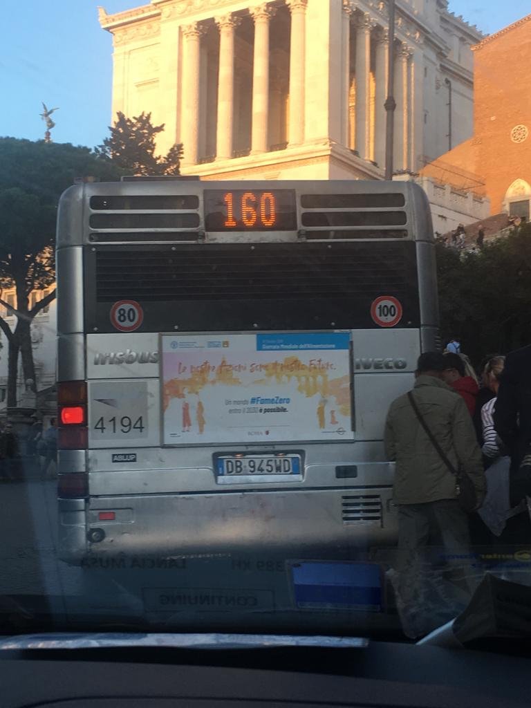 Rear view of a bus with a license plate DB 945WD, displaying a digital sign showing the number 160. The bus has various stickers, including a speed limit sign of 80 and 100. The background shows historic buildings and people walking.