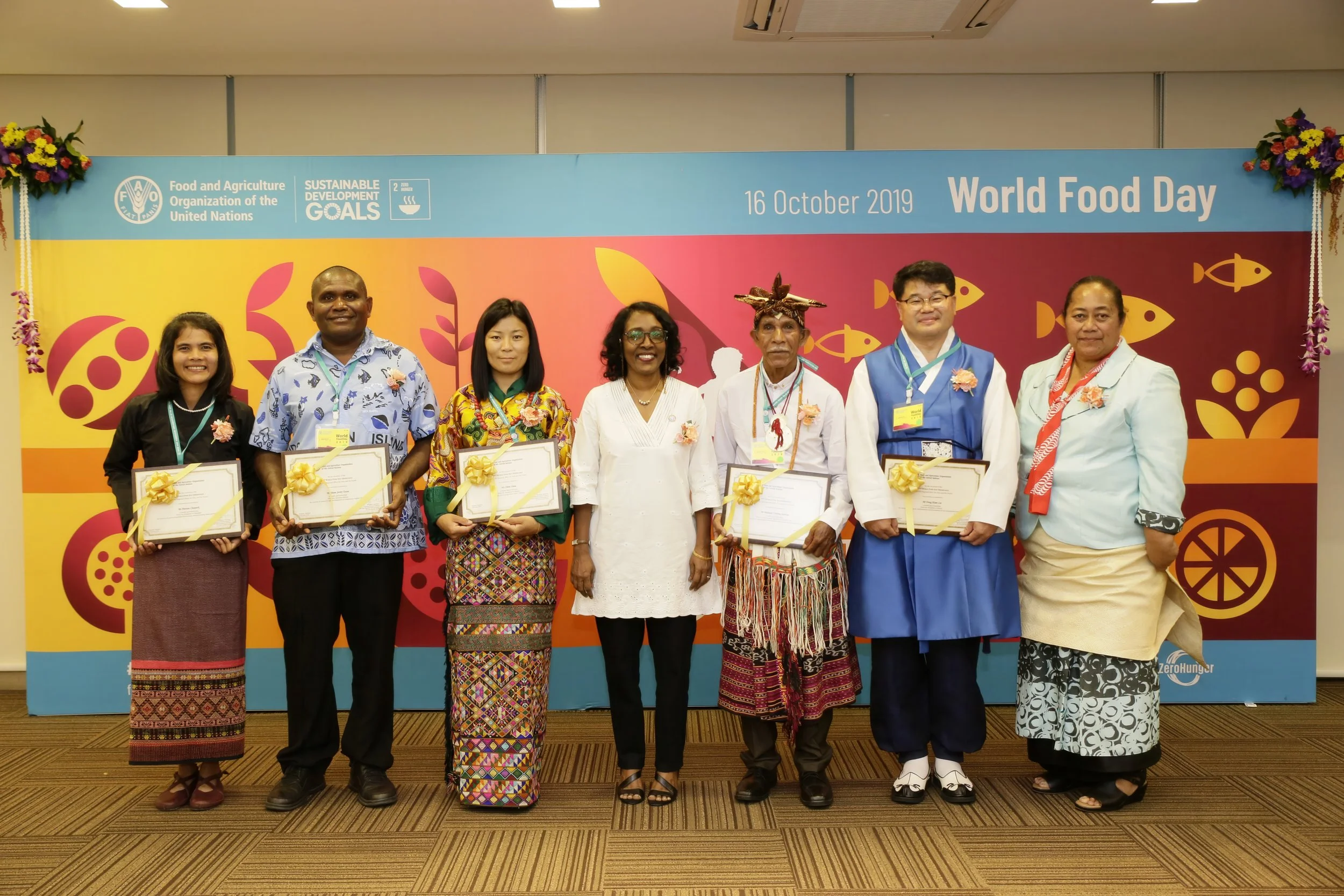 Group of eight diverse people standing in front of a colorful World Food Day banner, holding certificates and wearing floral decorations, at an indoor event.