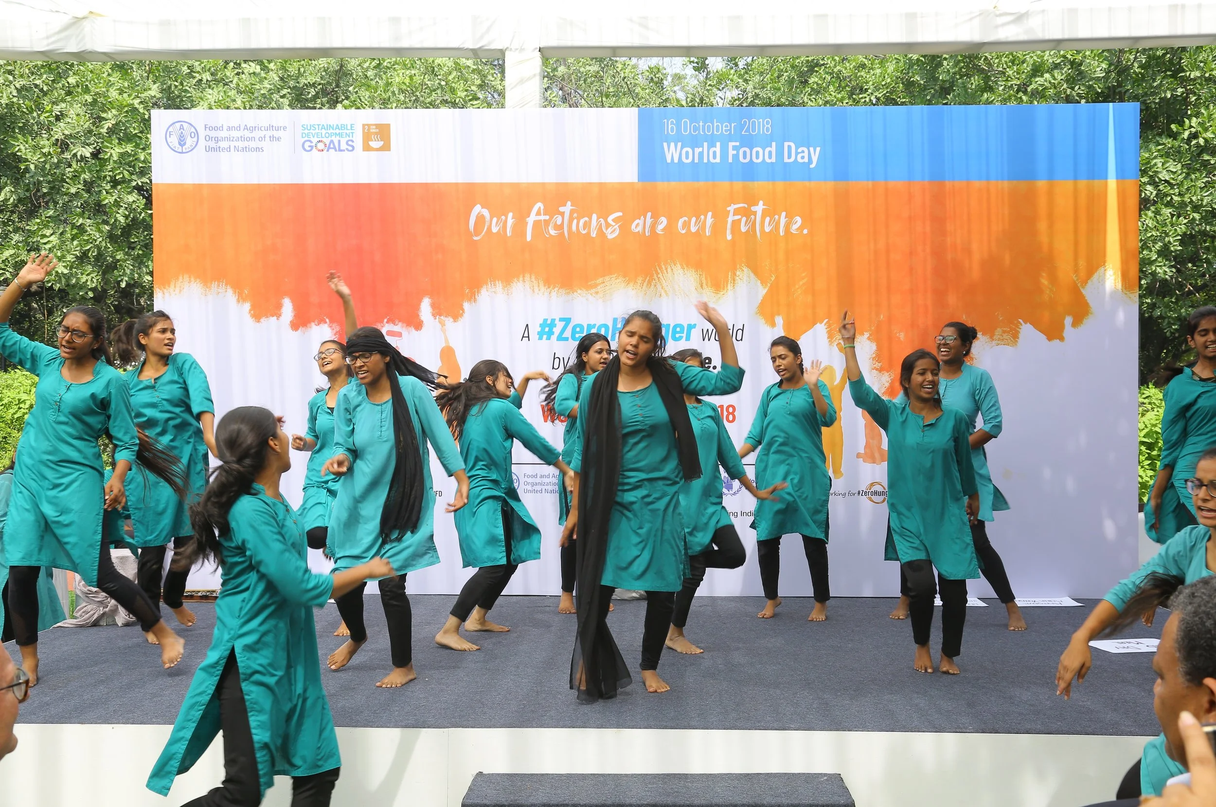 Group of women dancing on stage during an outdoor event for World Food Day, with a large banner in the background displaying event information and slogans.