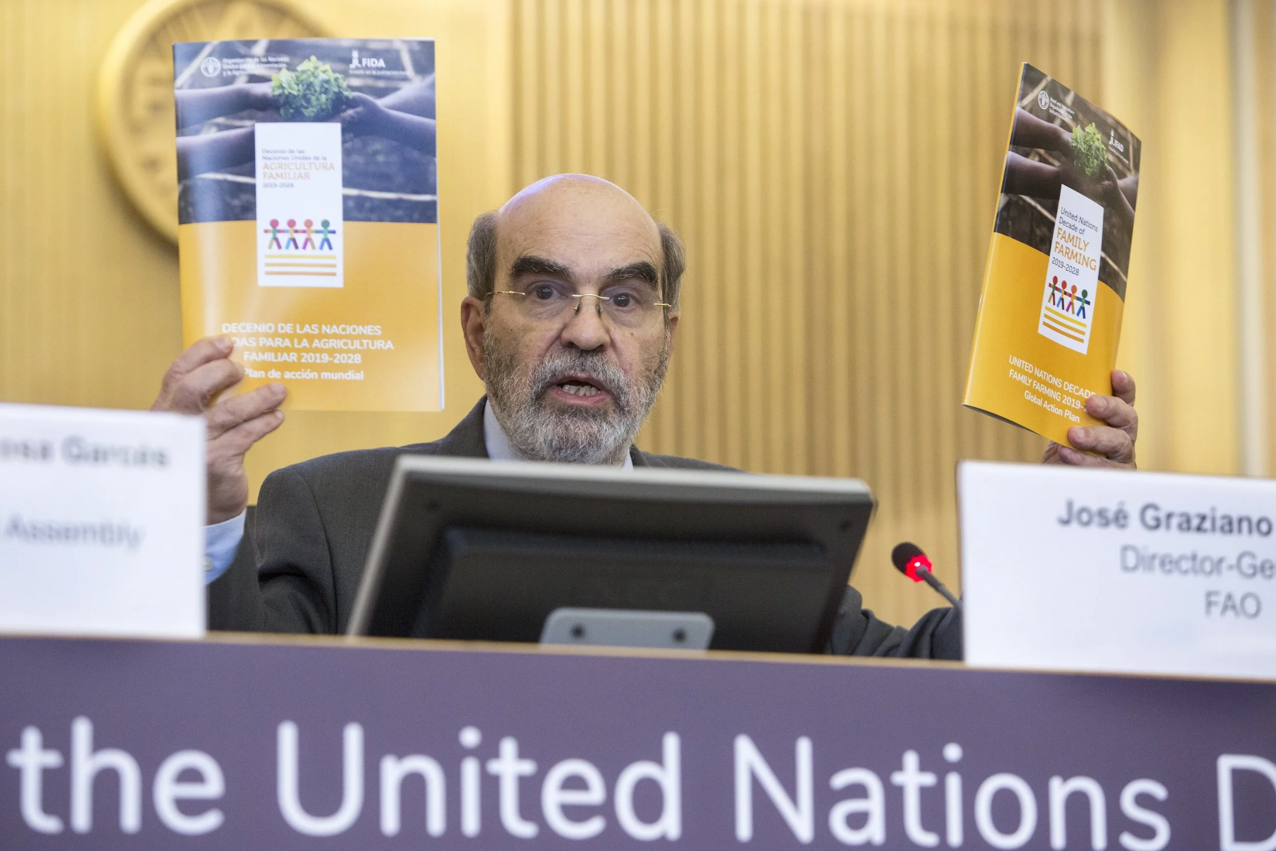 A man with glasses and a beard holds up two booklets titled 'United Nations Decade of Family Farming 2019-2028' during a conference. There are nameplates and a microphone in front of him, and a world map in the background.