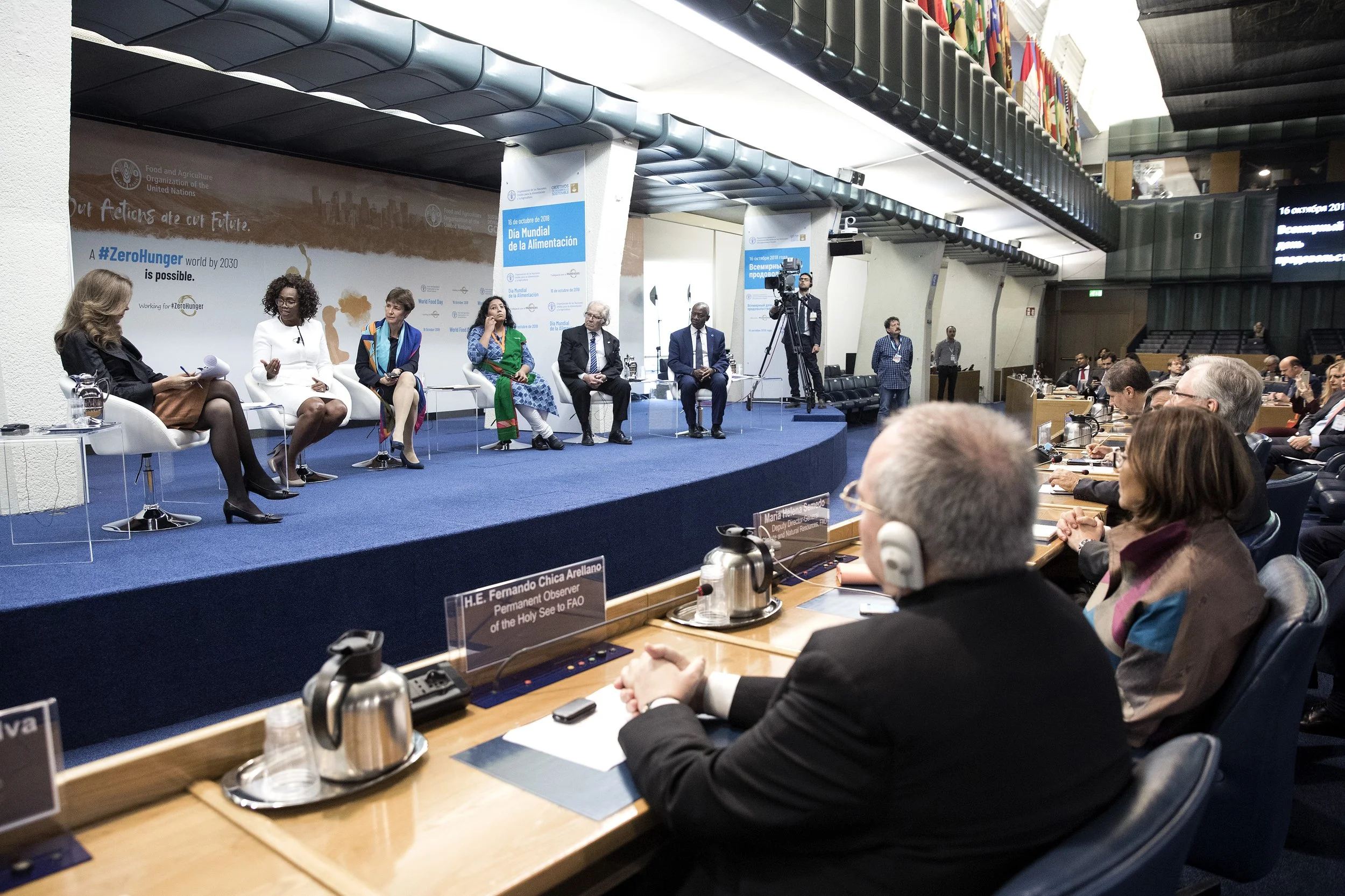 A panel of six diverse individuals seated on a stage in front of a large blue banner at a conference or event. The audience, mostly dressed in business attire, is seated and attentive. The room features flags on the upper level and tables with microp