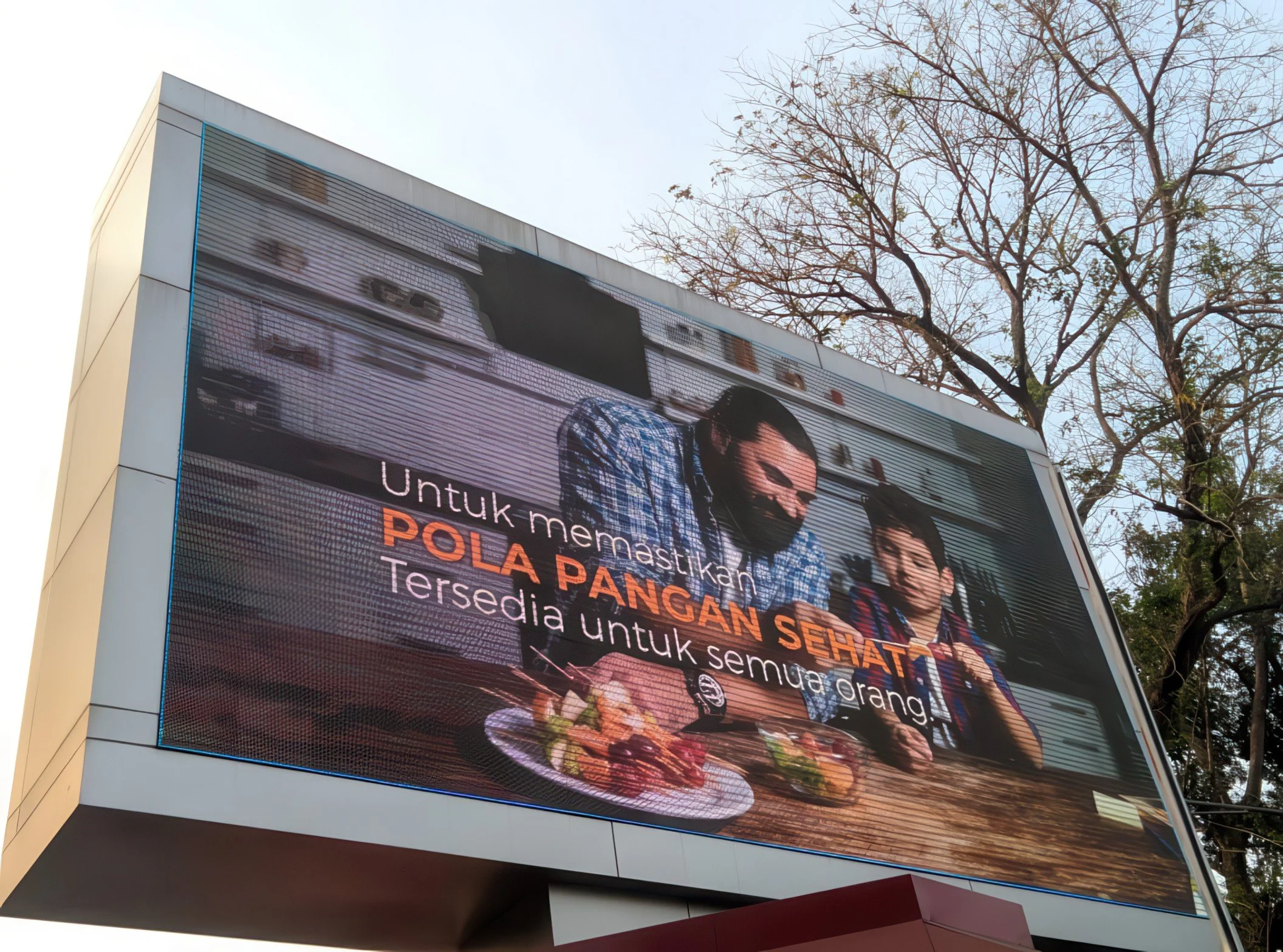 A large outdoor electronic billboard displaying an advertisement with the faces of three women. The billboard is mounted on a modern white building with a tree with bare branches nearby.